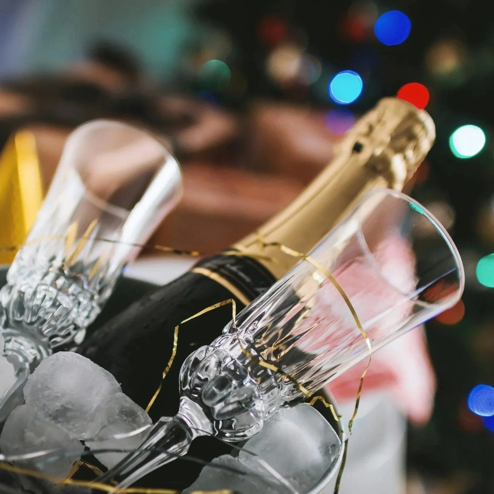 Close-up of a champagne bottle with a gold foil cap, surrounded by two champagne flutes with gold ribbons, all resting on ice cubes, with blurred colorful Christmas lights in the background.