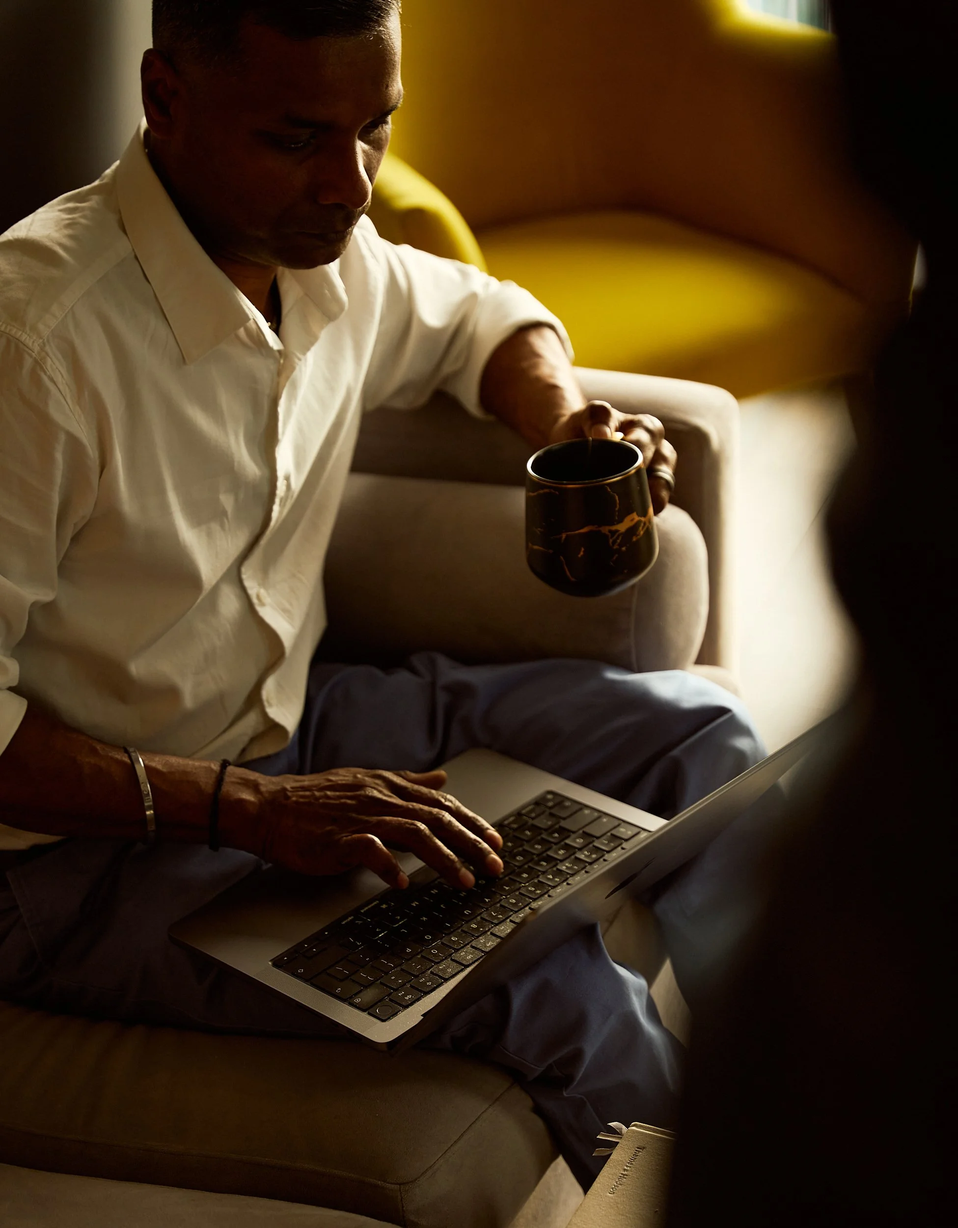A woman with long brown hair sitting on a yellow chair in a modern office or cafe, smiling and looking at her smartphone while holding a white mug.