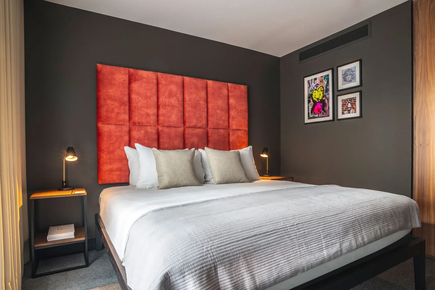 Modern bedroom with a large bed, orange upholstered headboard, dark gray walls, bedside lamps, and framed artwork.