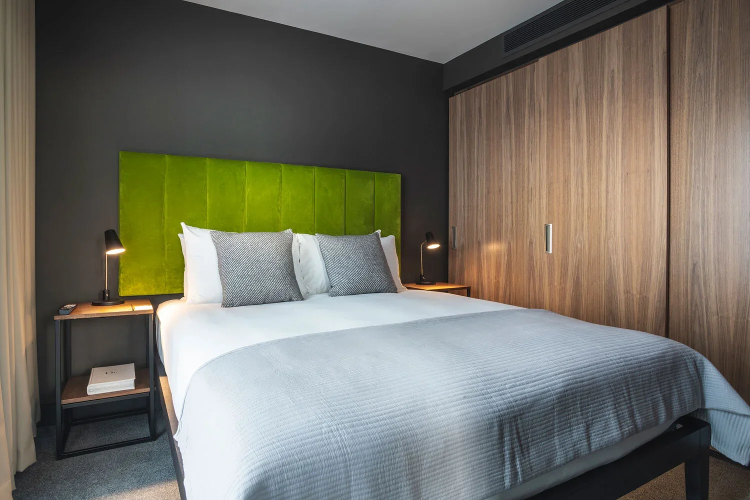 A neatly made bed with white linens, gray pillows, and a green upholstered headboard, flanked by two black bedside tables with black desk lamps in a modern hotel room with a dark accent wall and wooden closet doors.