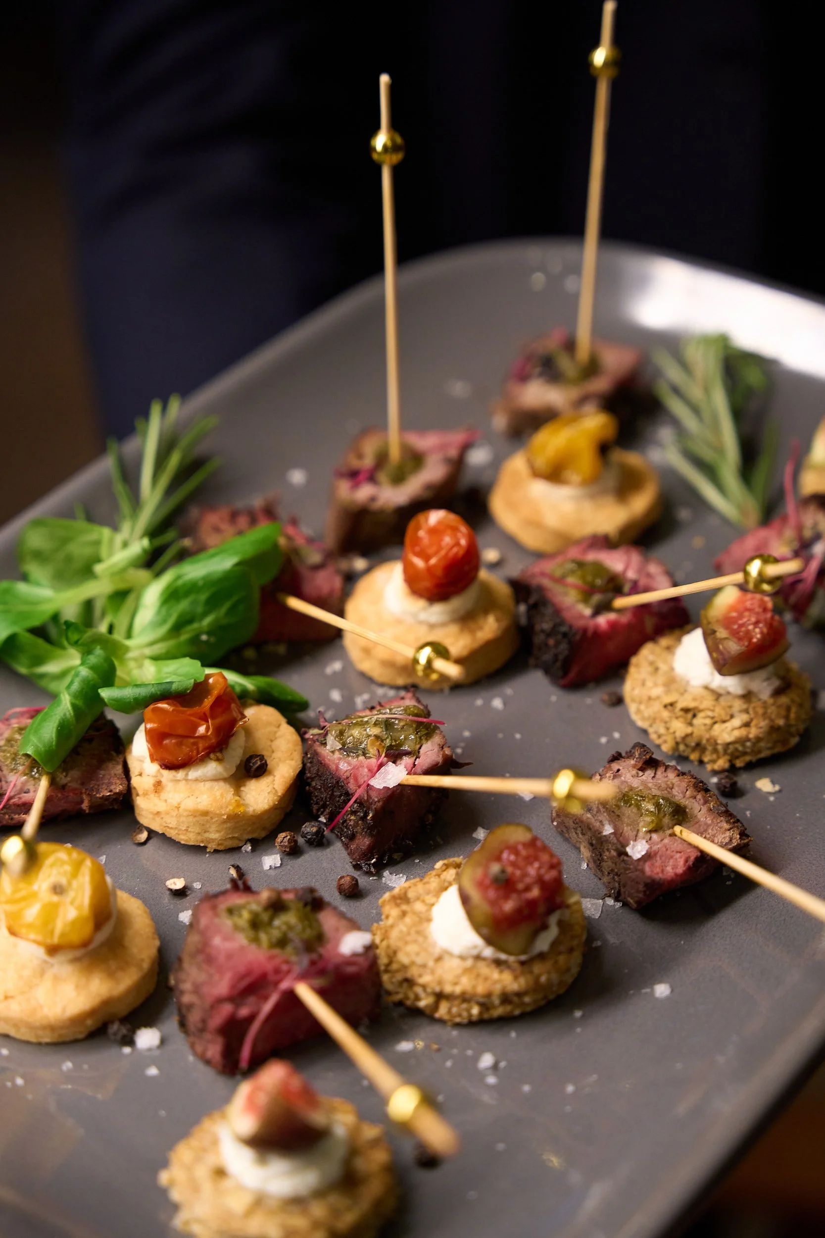 Assorted gourmet appetizers on a tray, featuring small bites with meats, cheeses, herbs, and garnishes, with decorative rosemary sprigs.