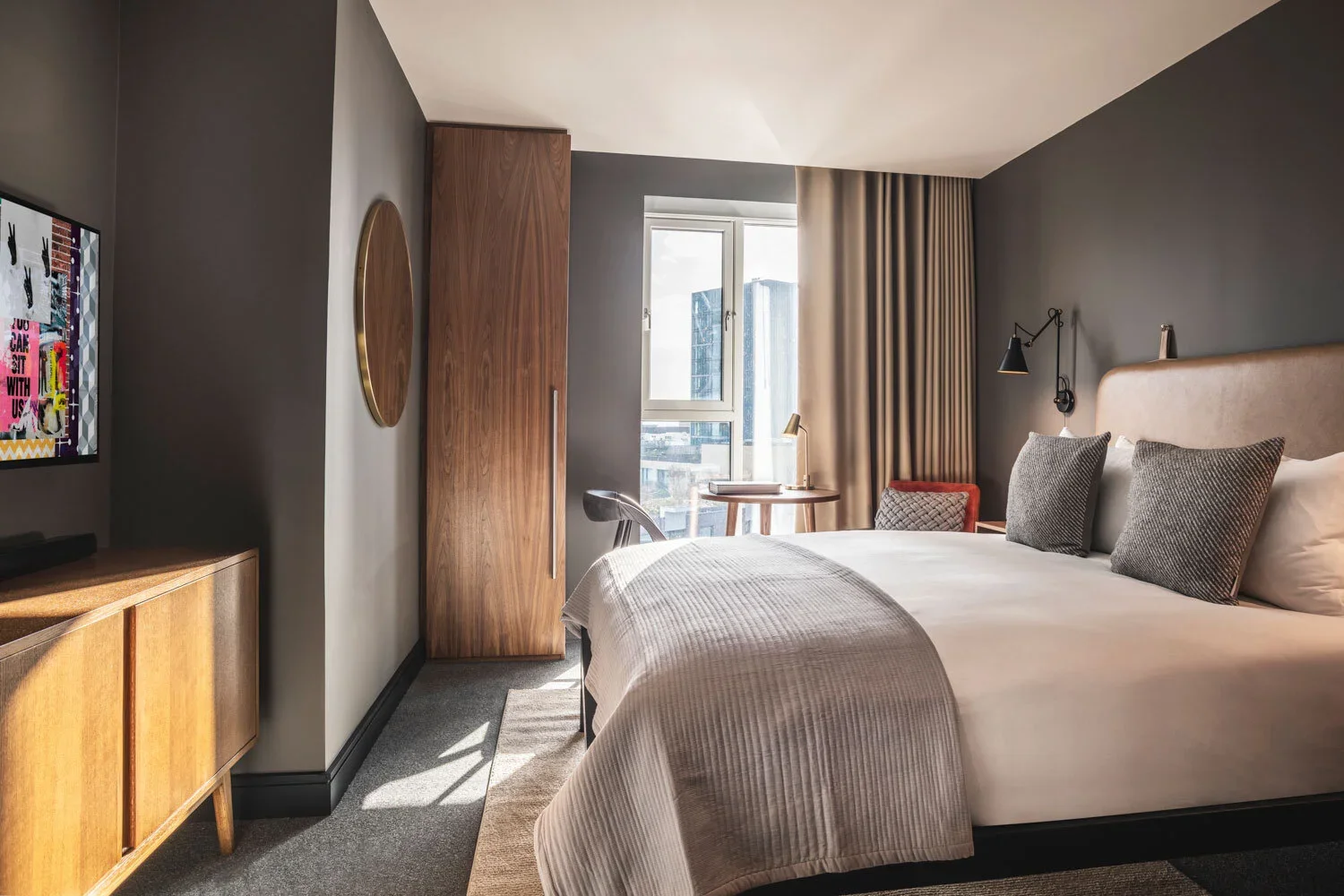 Modern hotel room with a large bed, gray walls, a wooden wardrobe, a window with beige curtains, a small round table with a desk lamp, and a TV on a wooden cabinet.