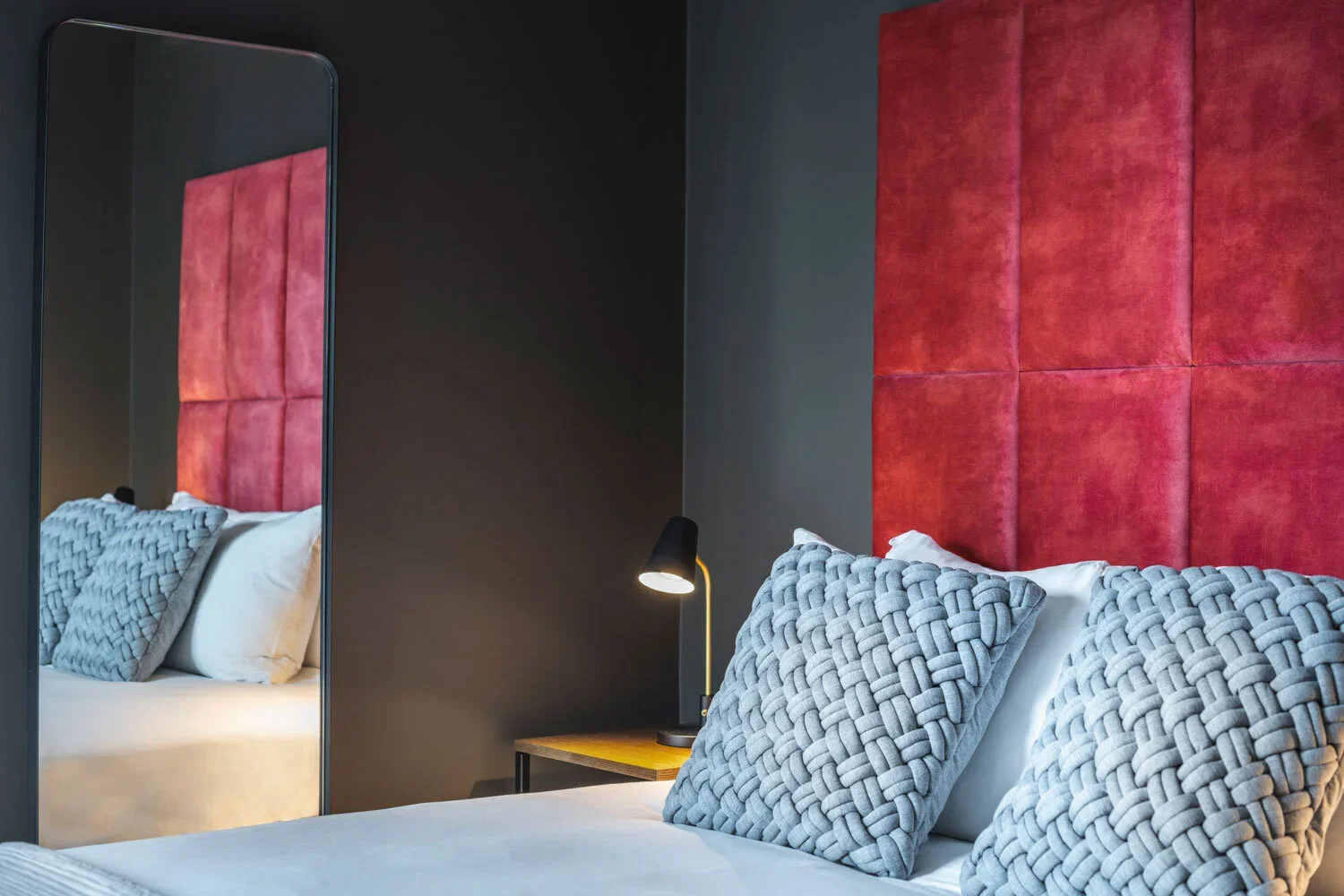 A bedroom with a bed, decorative pillows, a side table, and a red upholstered headboard, reflected in a mirror.