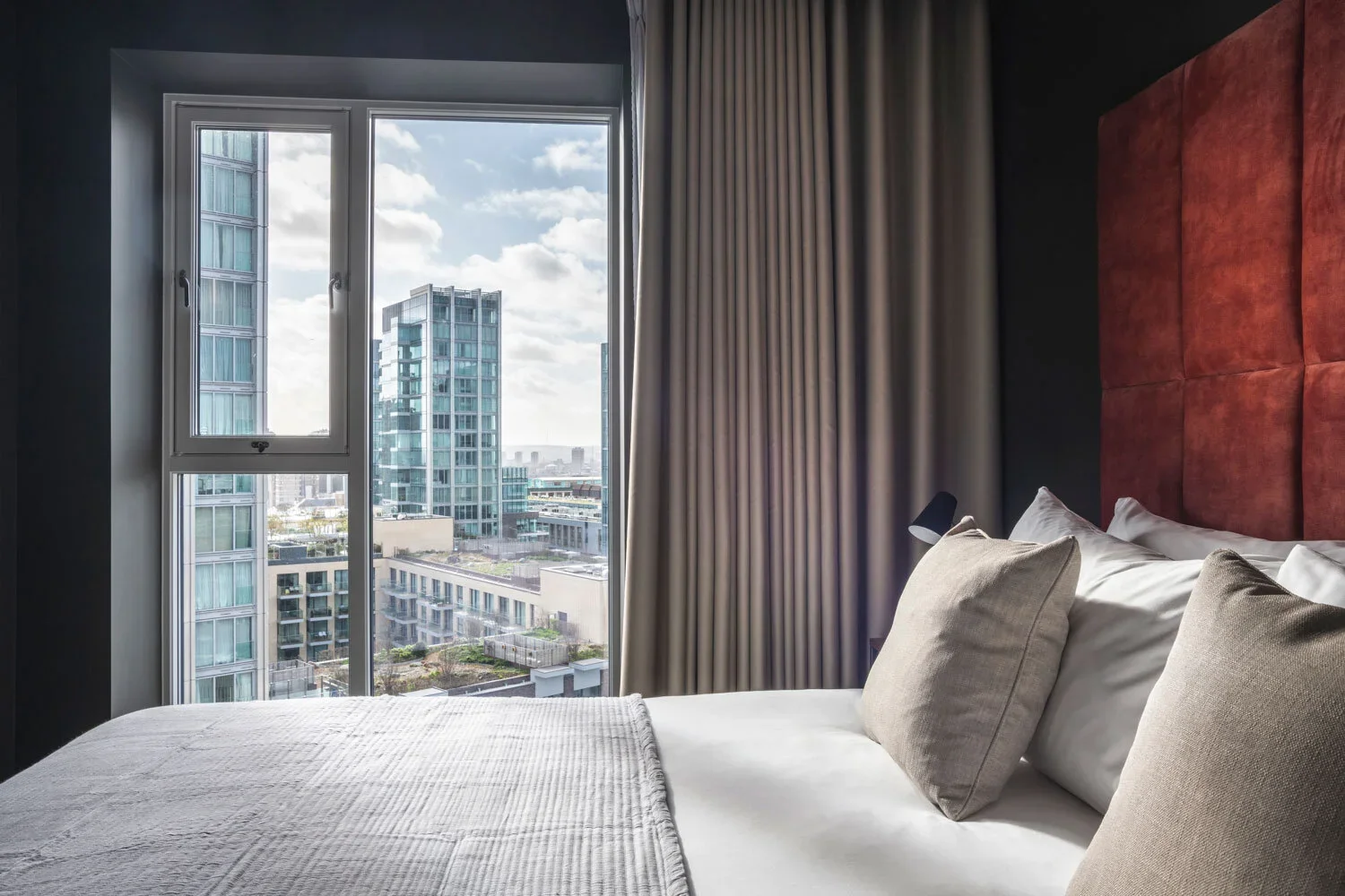 Modern hotel room with a bed, beige pillows, dark wall, large window showing city skyscrapers, and beige curtains.