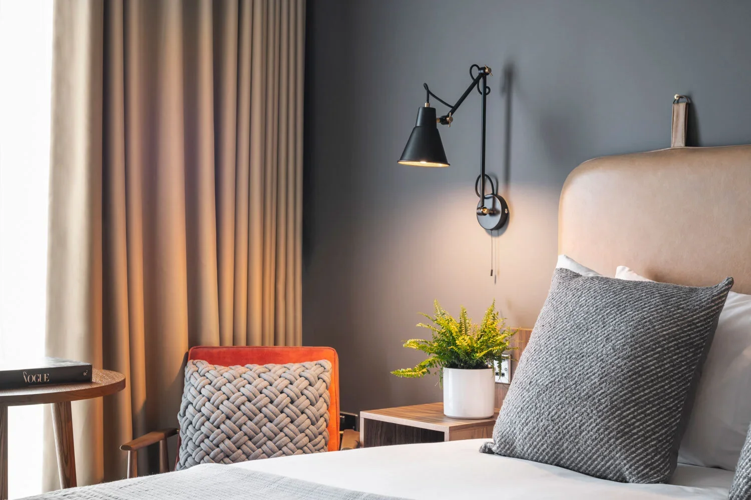 A cozy bedroom corner with a bed, gray pillows, a wooden nightstand with a potted plant, a chair with a decorative cushion, a window with beige curtains, and a black wall-mounted lamp.