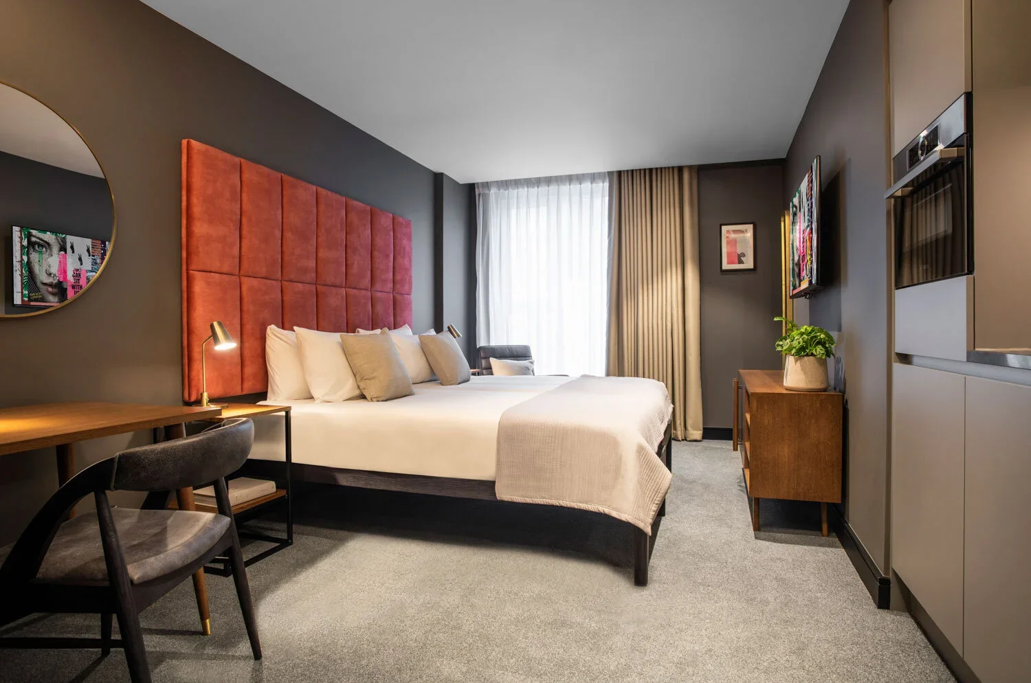 Modern hotel room with a large bed, pink cushioned headboard, decorative pillows, a wooden desk with a lamp, a gray chair, a small wooden side table with a plant, a wall-mounted TV, and window with curtains.