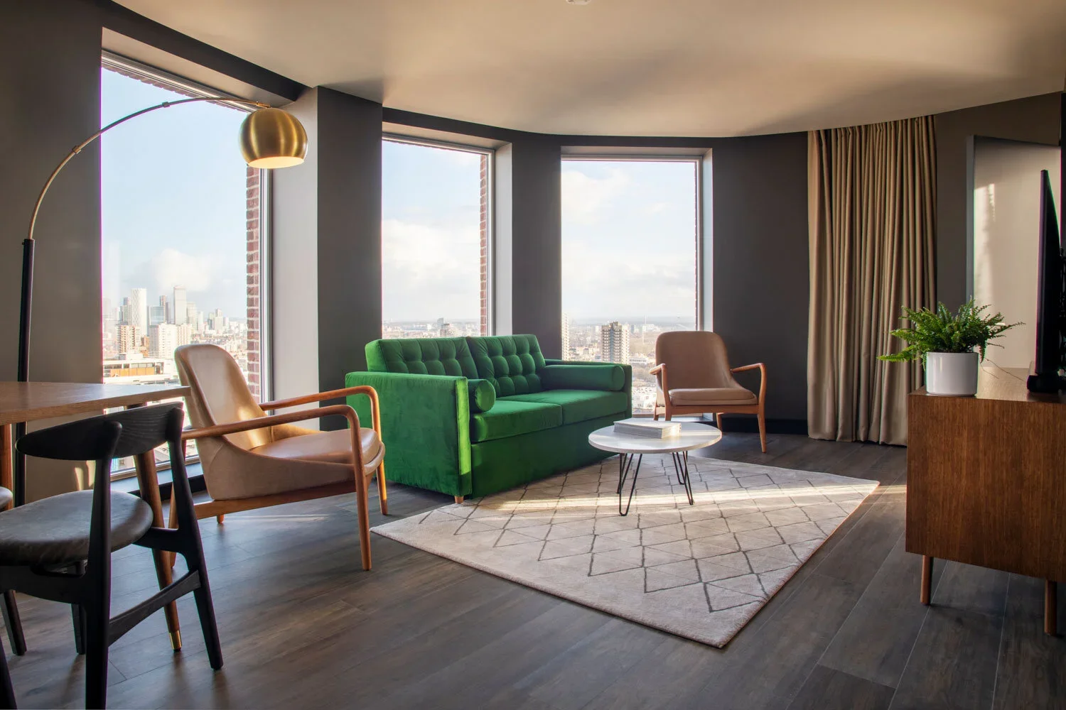Modern living room with large windows, green velvet sofa, beige armchairs, a round coffee table, wooden chairs, a floor lamp with a brass shade, a patterned area rug, potted plant, and curtains.