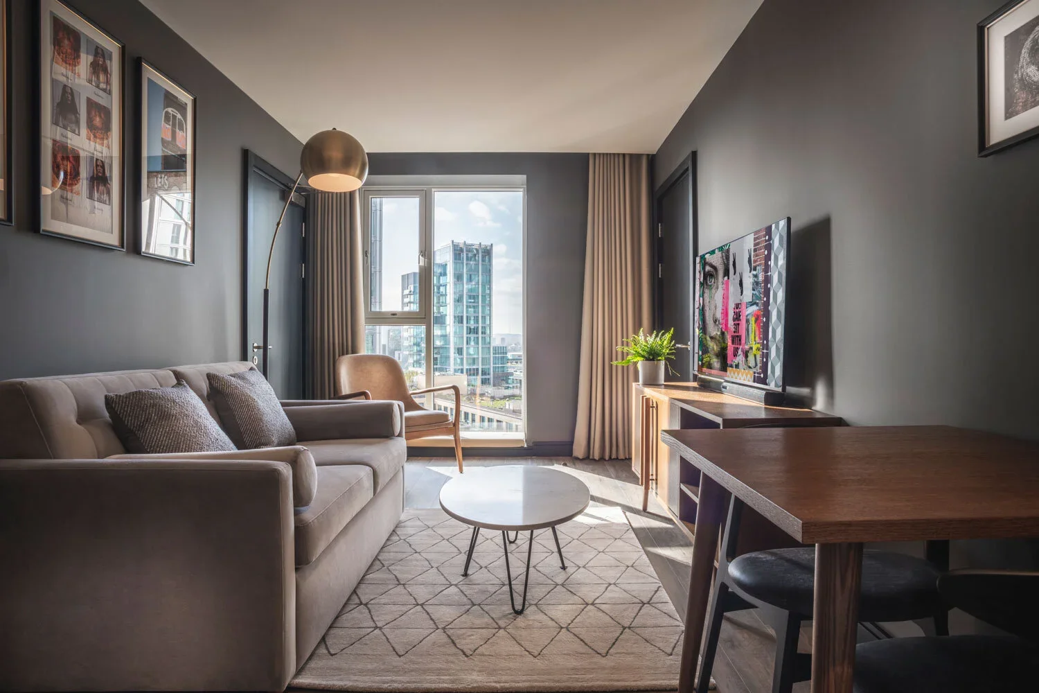 Modern living room with a beige sofa, coffee table, armchair, television, potted plant, wall art, large window with city view, and beige curtains.