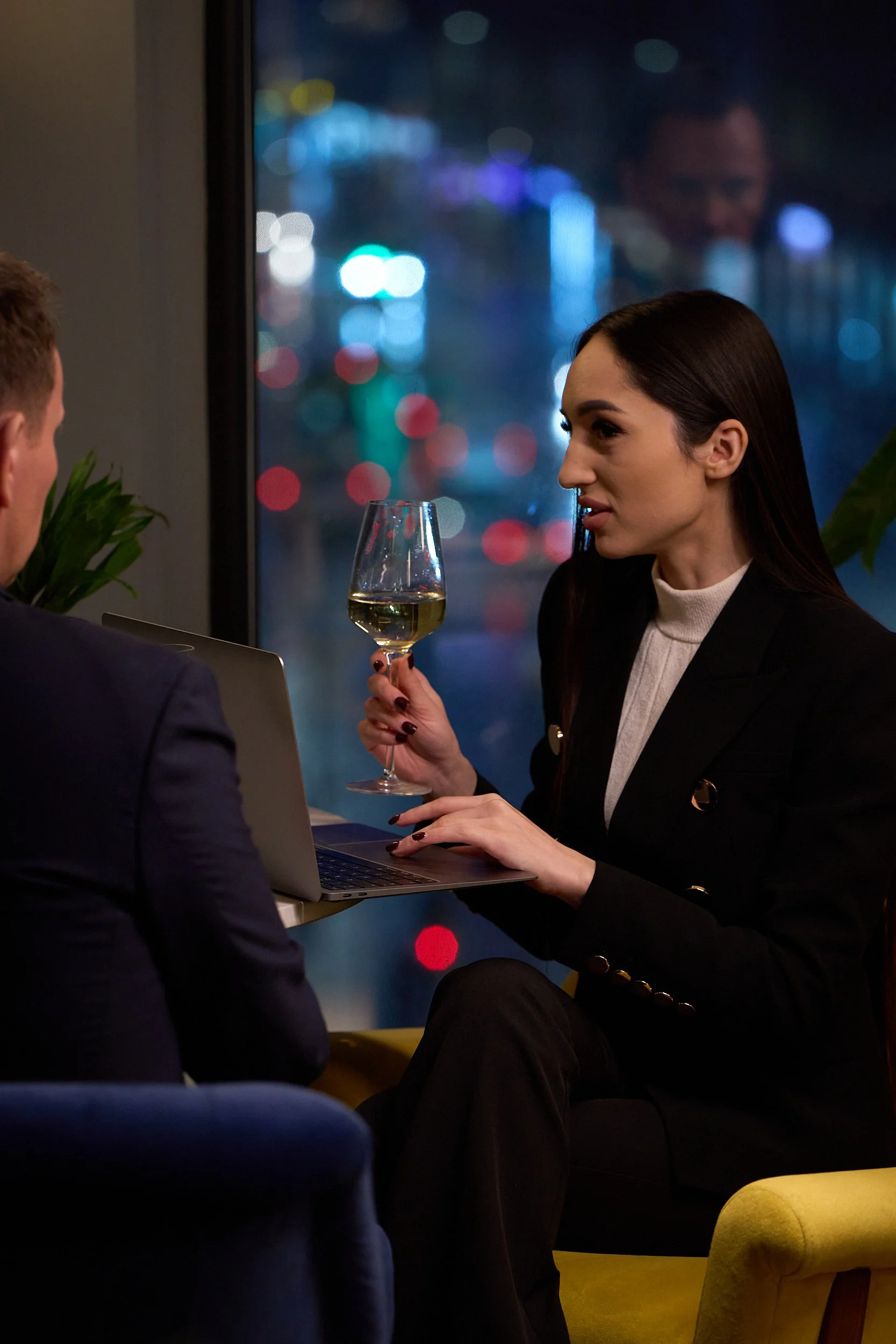 A woman with dark hair wearing a black blazer and white turtleneck, holding a glass of white wine, sitting across from a man with short hair, in a high-rise city setting with colorful city lights outside the window.