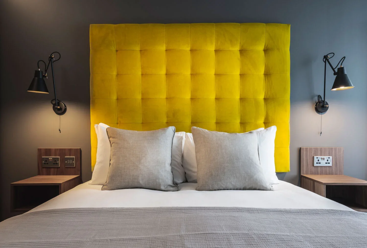 A modern bedroom with a large yellow tufted headboard, white bedding, gray pillows, two wall-mounted black reading lamps, and small wooden nightstands with electrical outlets.