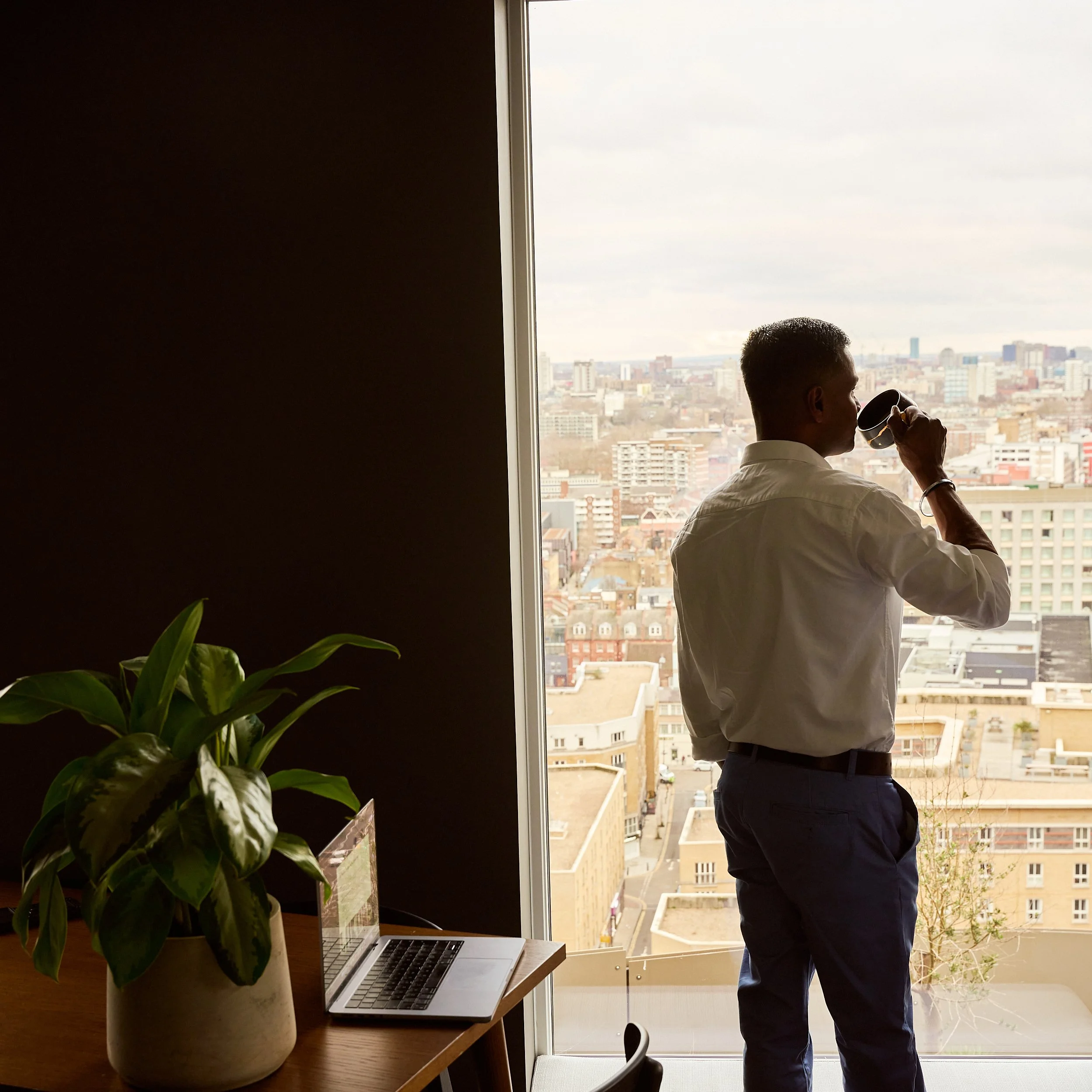 A man in formal attire stands near a large window looking out at a cityscape while drinking from a mug. On a nearby wooden table, there is a large potted green plant and an open laptop.