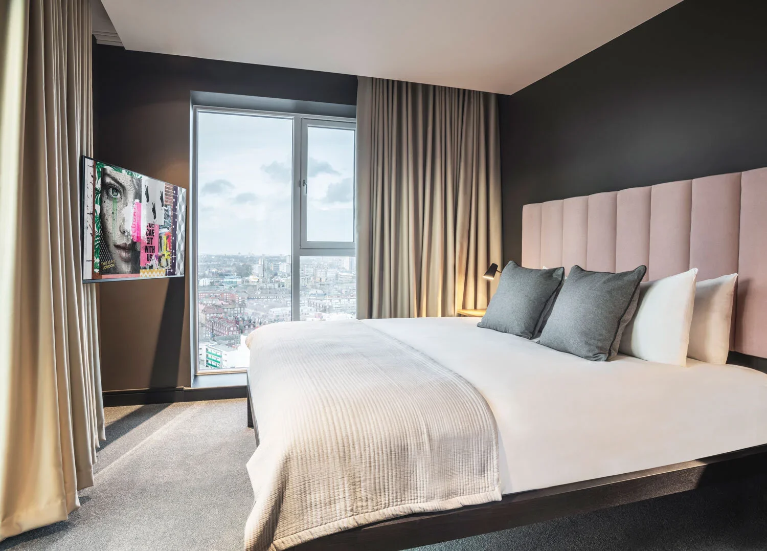Bedroom with large window, bed with pink headboard, gray pillows, white bedding, and mounted TV showing street view.