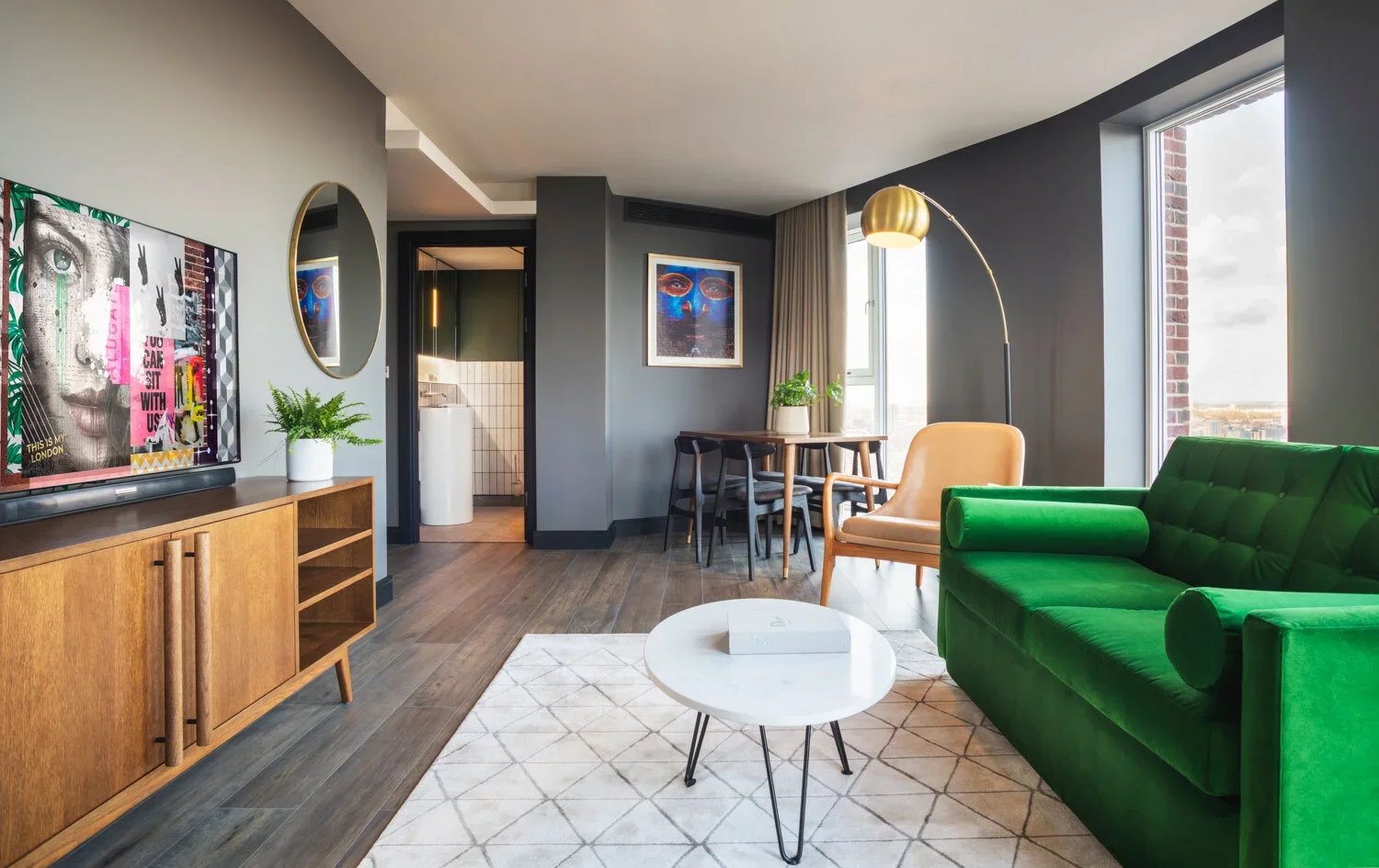 Modern living room with green velvet sofa, white round coffee table, and wooden sideboard, with artwork and plants, overlooking city through large window.