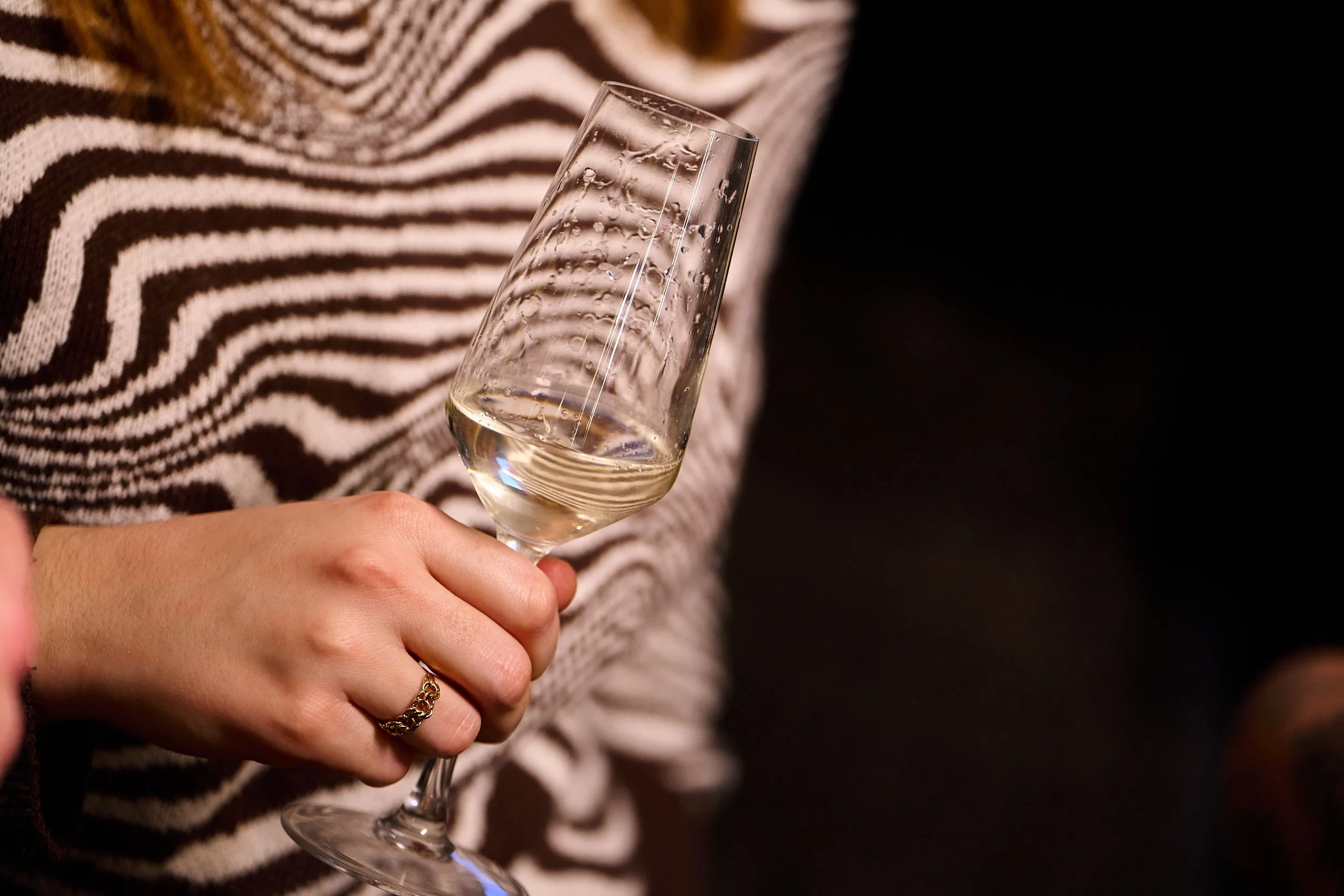 A person holding a champagne flute with bubbles inside, wearing a gold bracelet and a shirt with black and white wavy stripes.