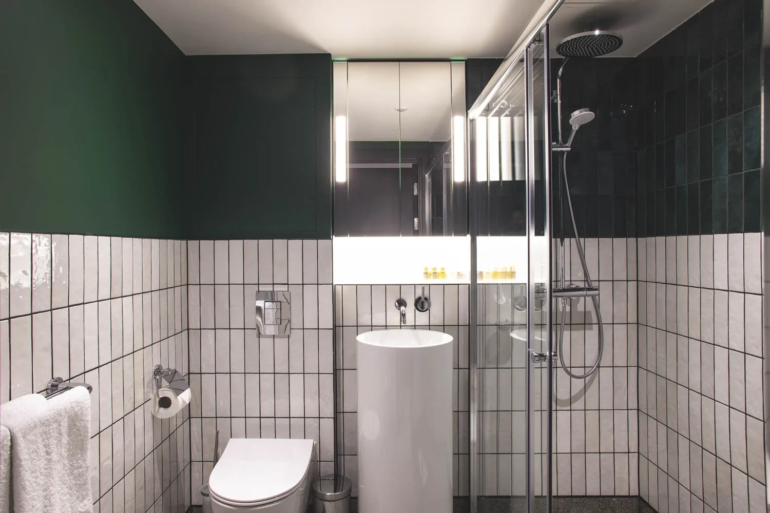 Modern bathroom with white tiled walls, dark green upper walls, a toilet, a pedestal sink, and a glass shower enclosure with a rain shower head, illuminated by vertical lights.