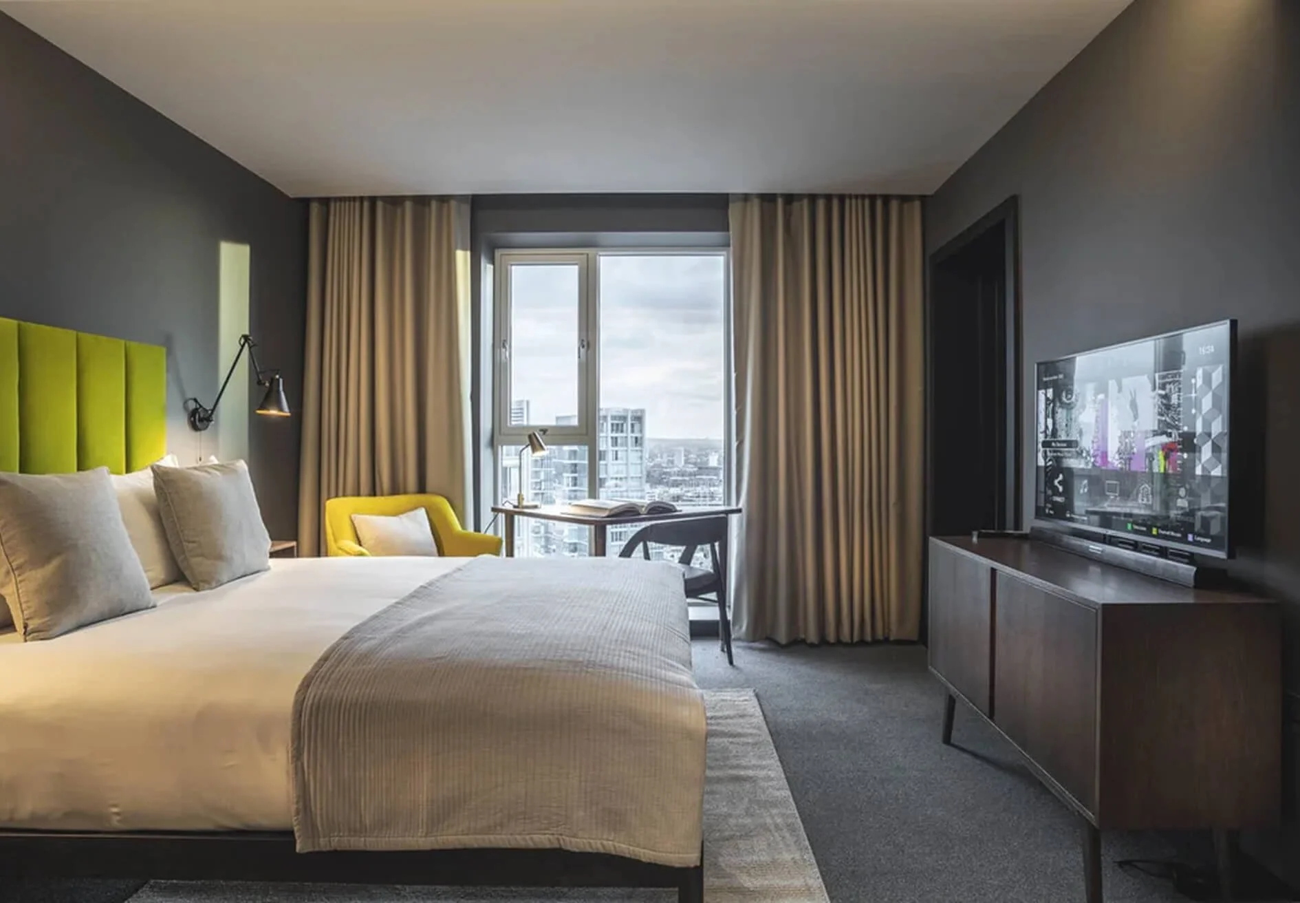 Modern hotel room with a bed, yellow accent chair, desk, and a city view through the window.
