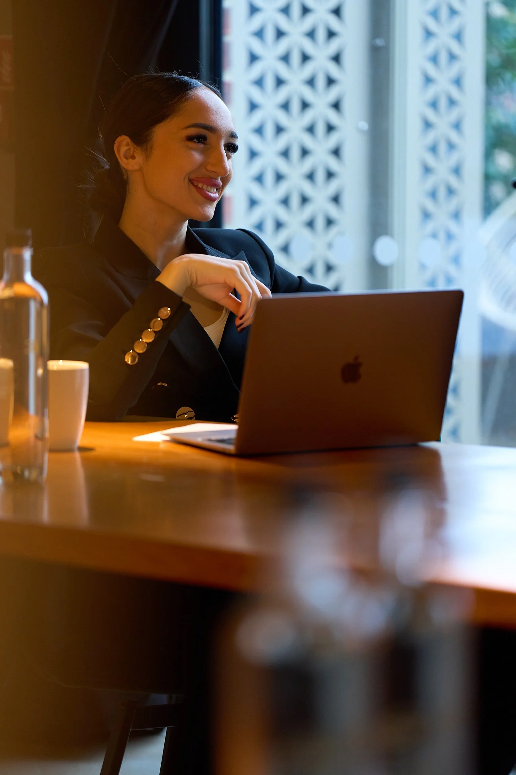 A woman with dark hair smiling while working on a laptop at a wooden table in a café or restaurant setting.