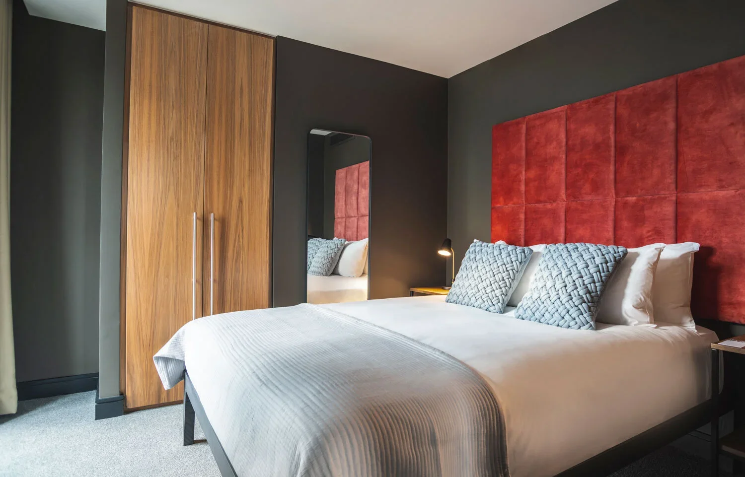 Modern bedroom with a large bed featuring a red upholstered headboard, white bedding, and decorative pillows, a wooden wardrobe, a mirror, and a bedside lamp.