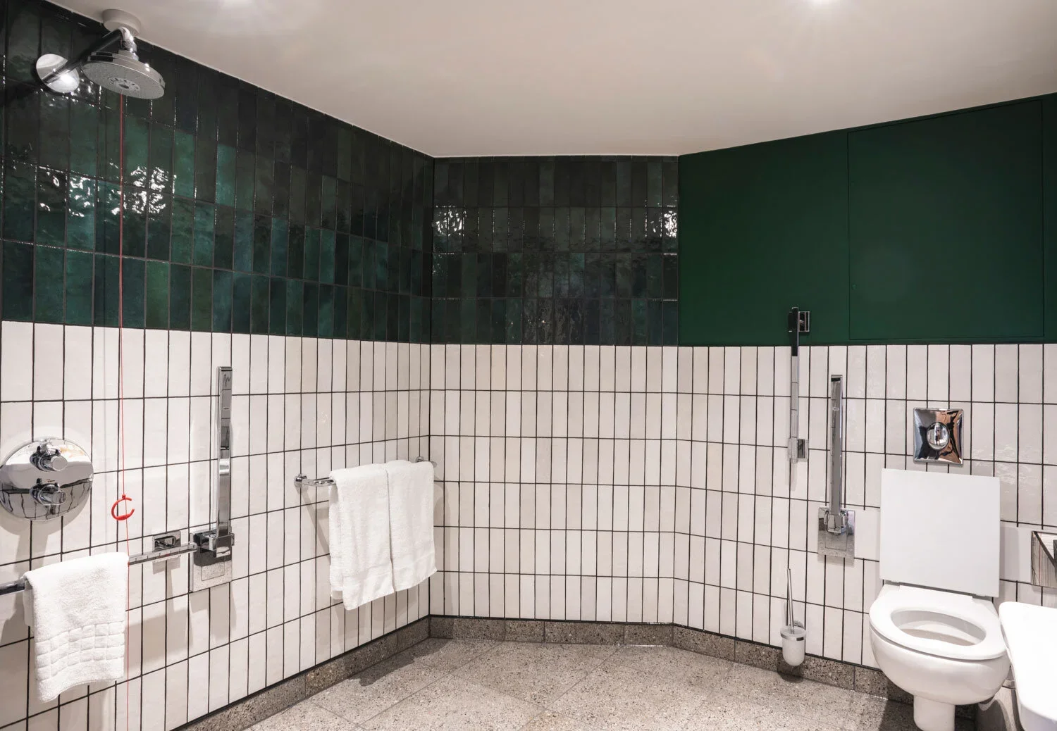 Accessible bathroom with green and black tiled walls, white tiled lower walls, a toilet, handrails, a showerhead, and white towels on a towel rack.