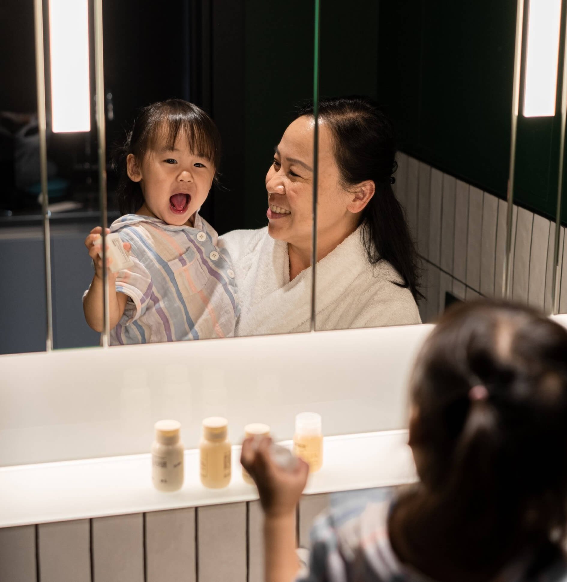 A woman and a young girl are looking at each other in front of a mirror in a bathroom. The girl is smiling with her mouth open, and the woman is smiling back.