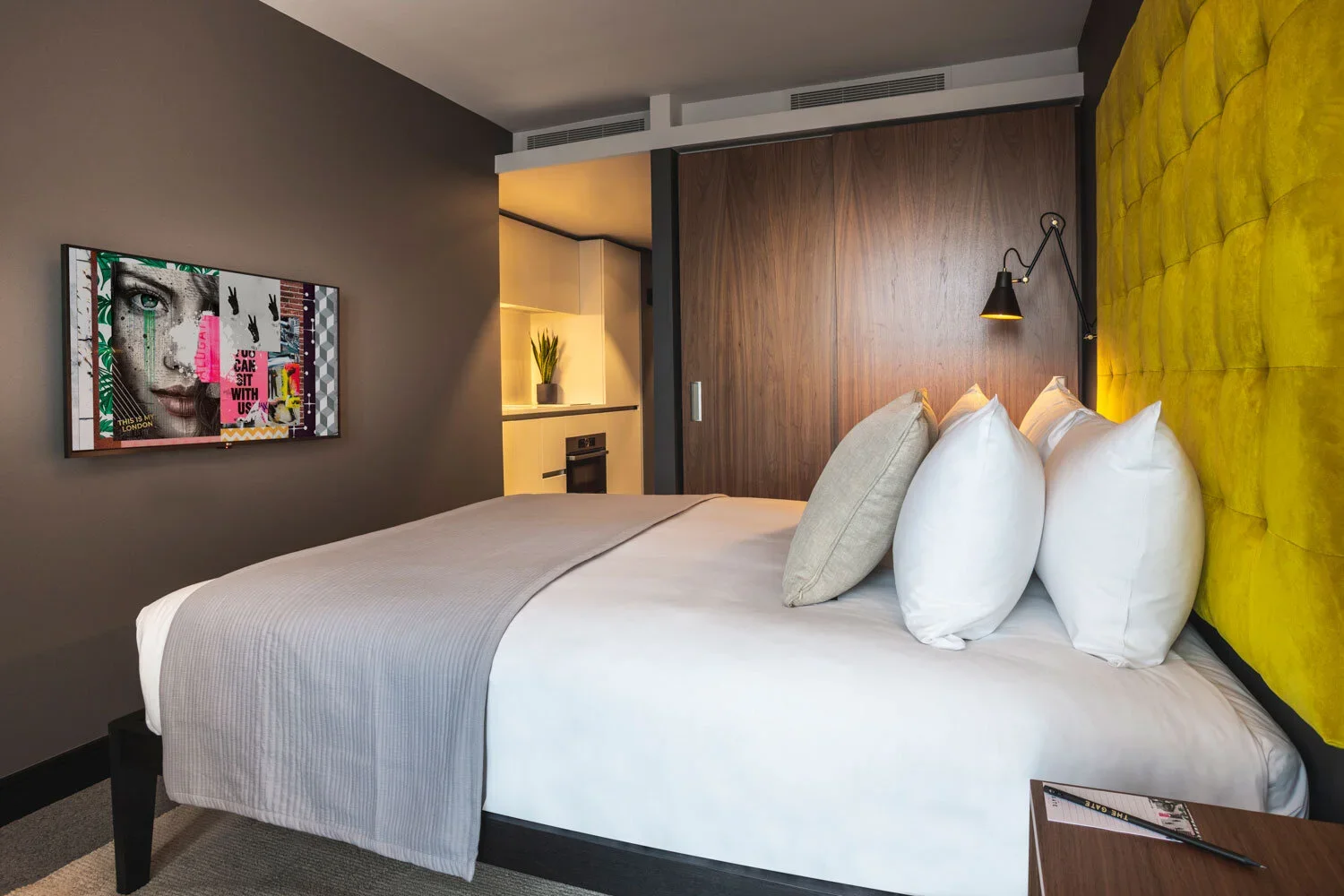 Modern hotel room with a large bed that has white sheets, beige pillows, and a yellow headboard, a wall-mounted black reading lamp, a colorful artwork on the wall, and a small kitchenette with a plant on the counter.