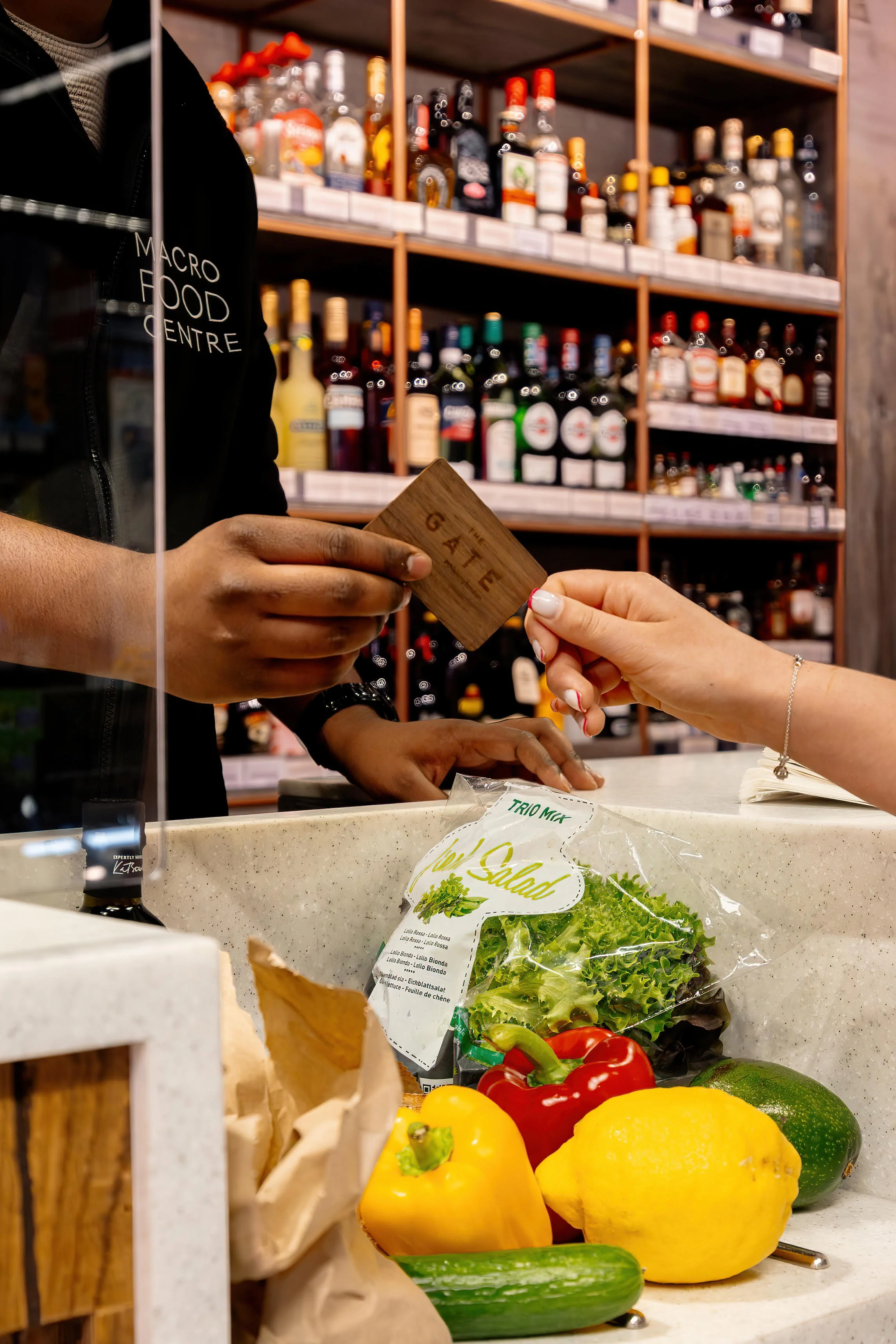 Customer at the grocery store checkout handing a loyalty card to cashier. Fresh vegetables, including yellow bell pepper, red bell peppers, lemon, and cucumber, are in the foreground. Shelves in the background are stocked with various bottles and jars.