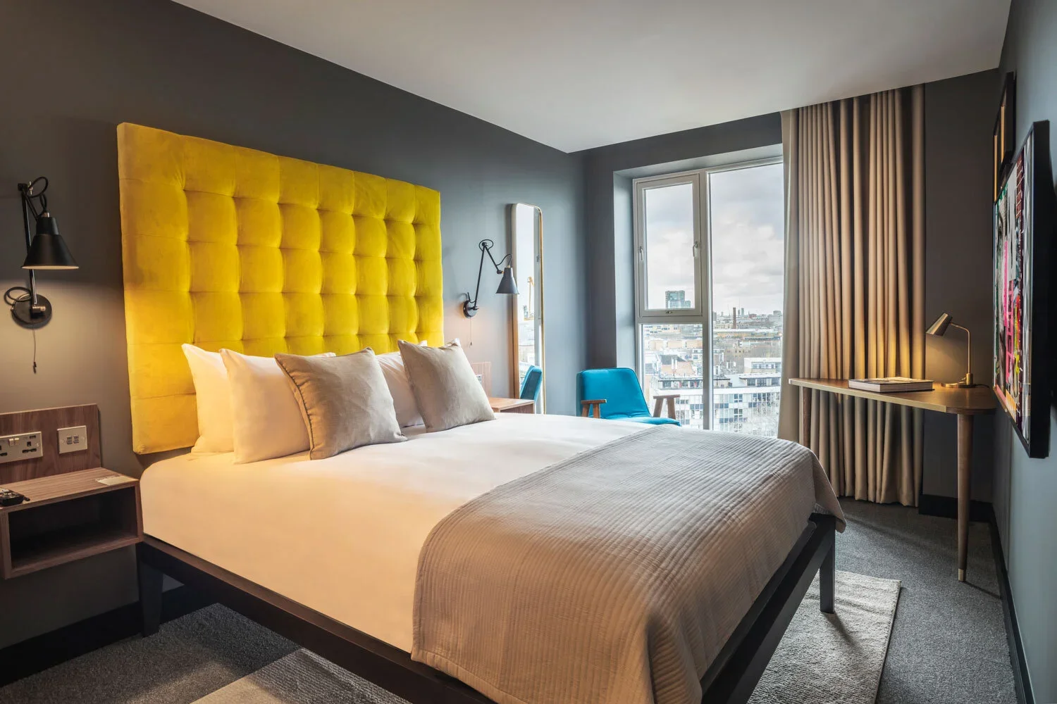 Hotel room with a large bed featuring a bright yellow, tufted headboard, gray walls, two black wall-mounted lamps, a nightstand with outlets, a blue armchair, a large window with city view, and a flat-screen TV.