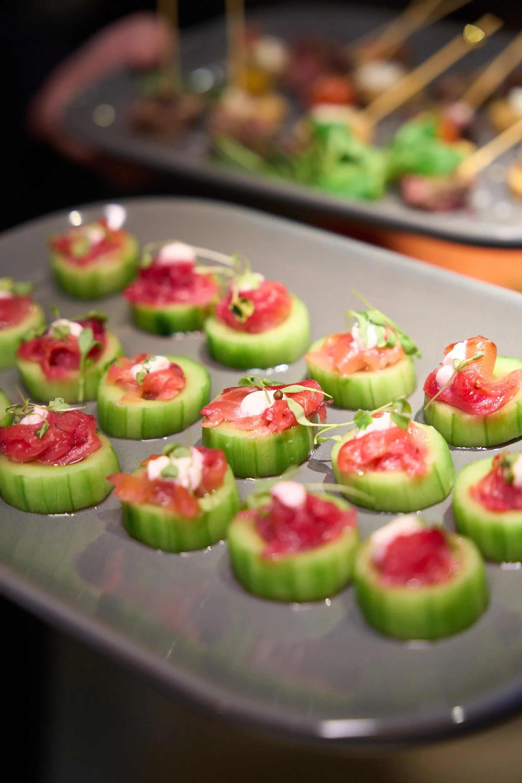 Close-up of cucumber slices topped with raw fish, possibly salmon, garnished with microgreens on a tray, with more appetizers in the background.