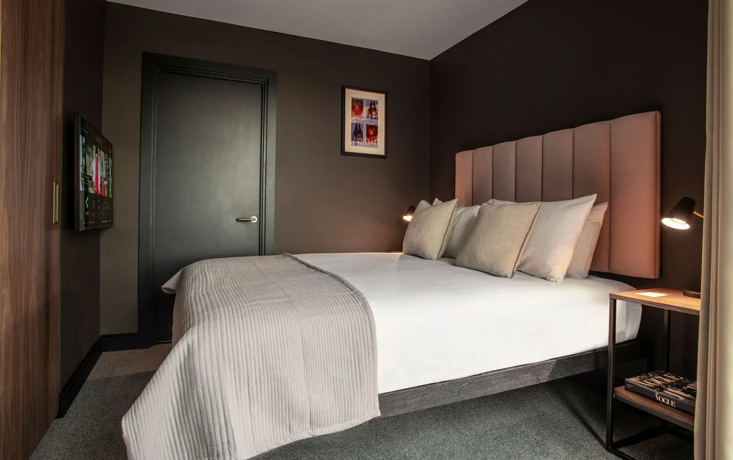 Modern hotel room with a bed, beige headboard, white bedding, beige pillows, dark walls, a small wooden nightstand with a lamp and magazines, and a TV mounted on the wall.