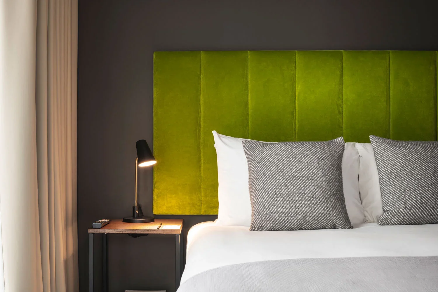 A hotel bed with white and gray bedding, a green velvet headboard, and a nightstand with a black lamp and remote control.