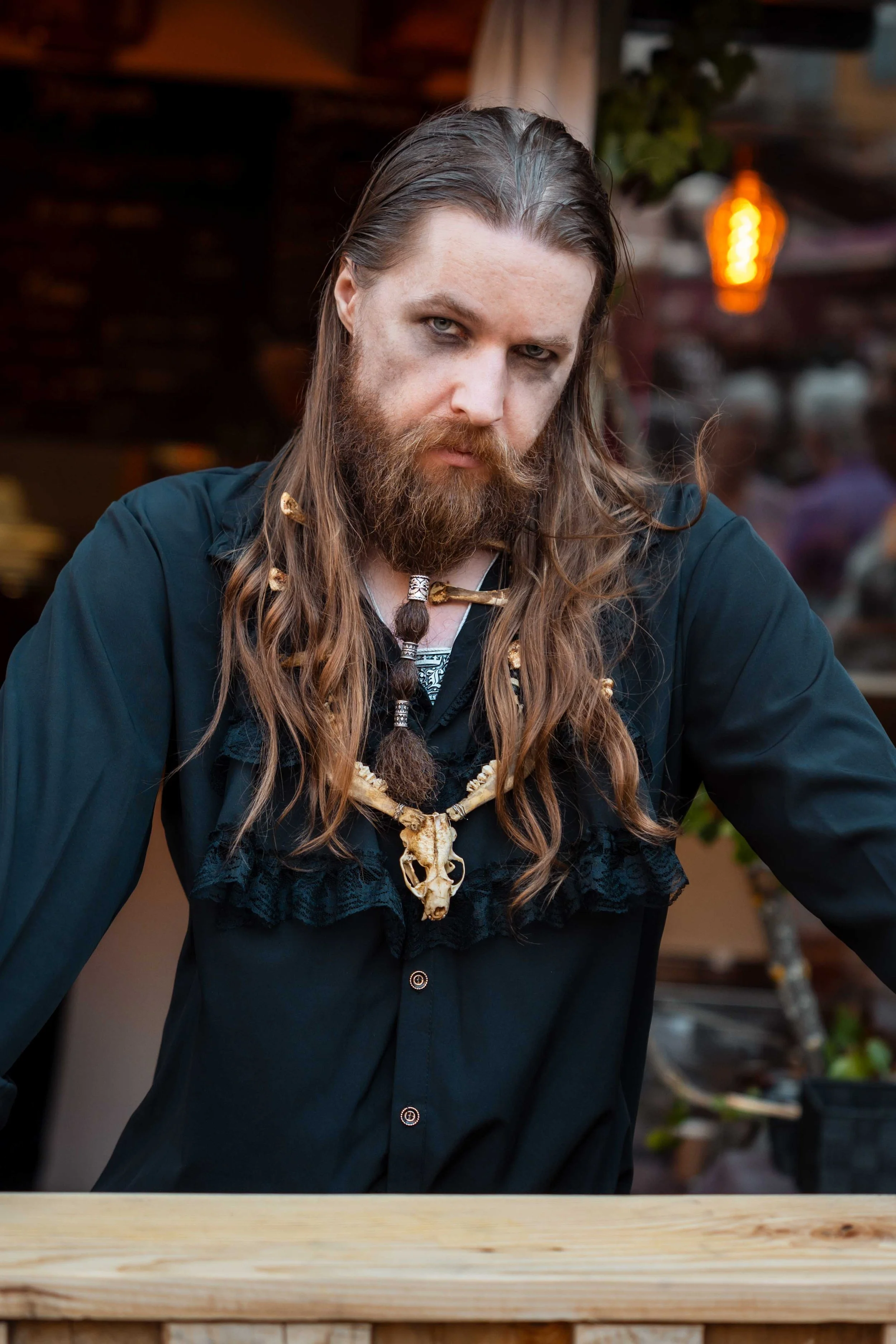 Homme avec longue chevelure et barbe, portant un collier en os et un vêtement noir, regardant fixement vers l'objectif, dans un cadre intérieur avec ambiance chaleureuse.
