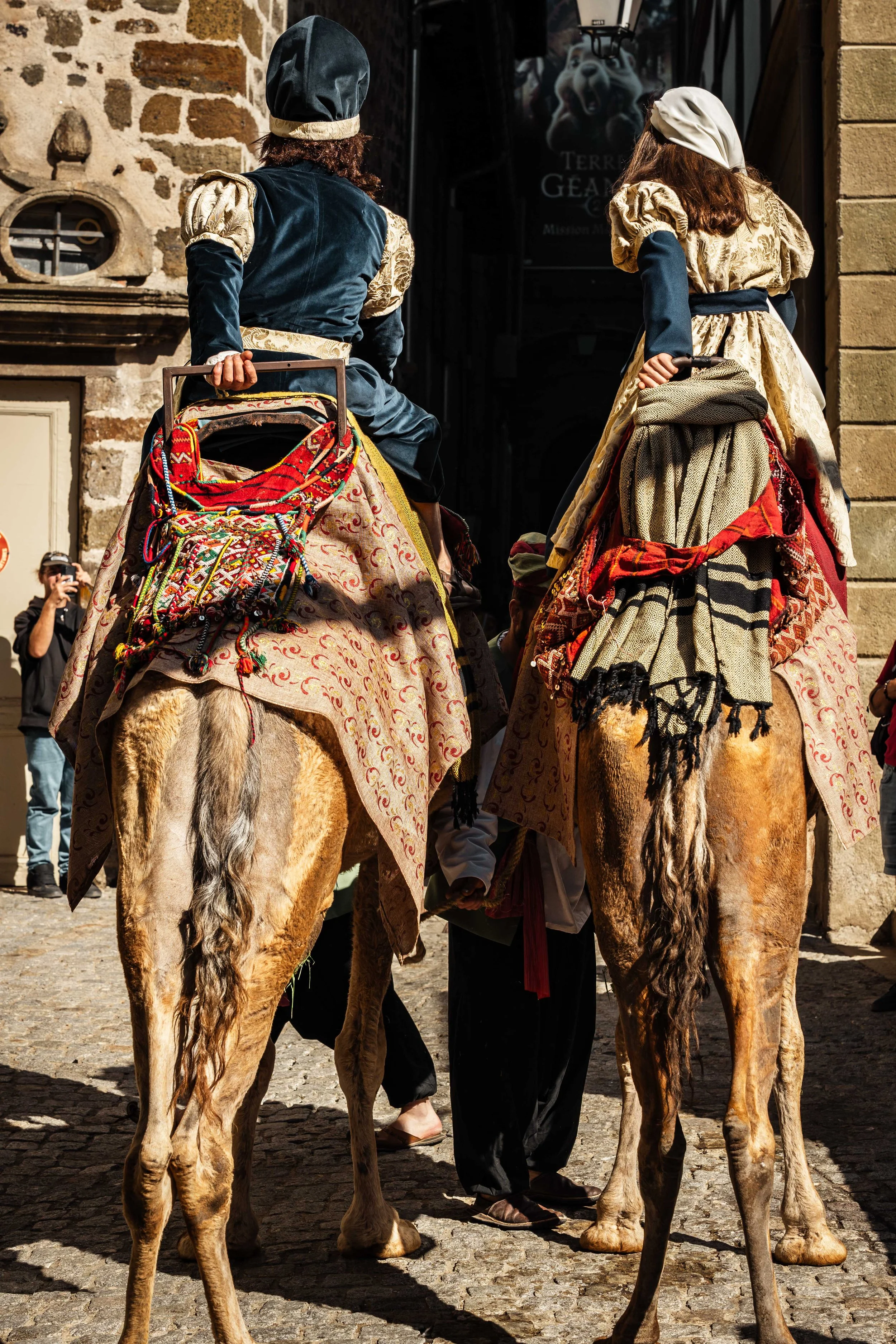 Deux personnes habillées en costumes traditionnels, assises à dos de chameaux, dans une rue pavée, avec plusieurs personnes et bâtiments anciens en arrière-plan.