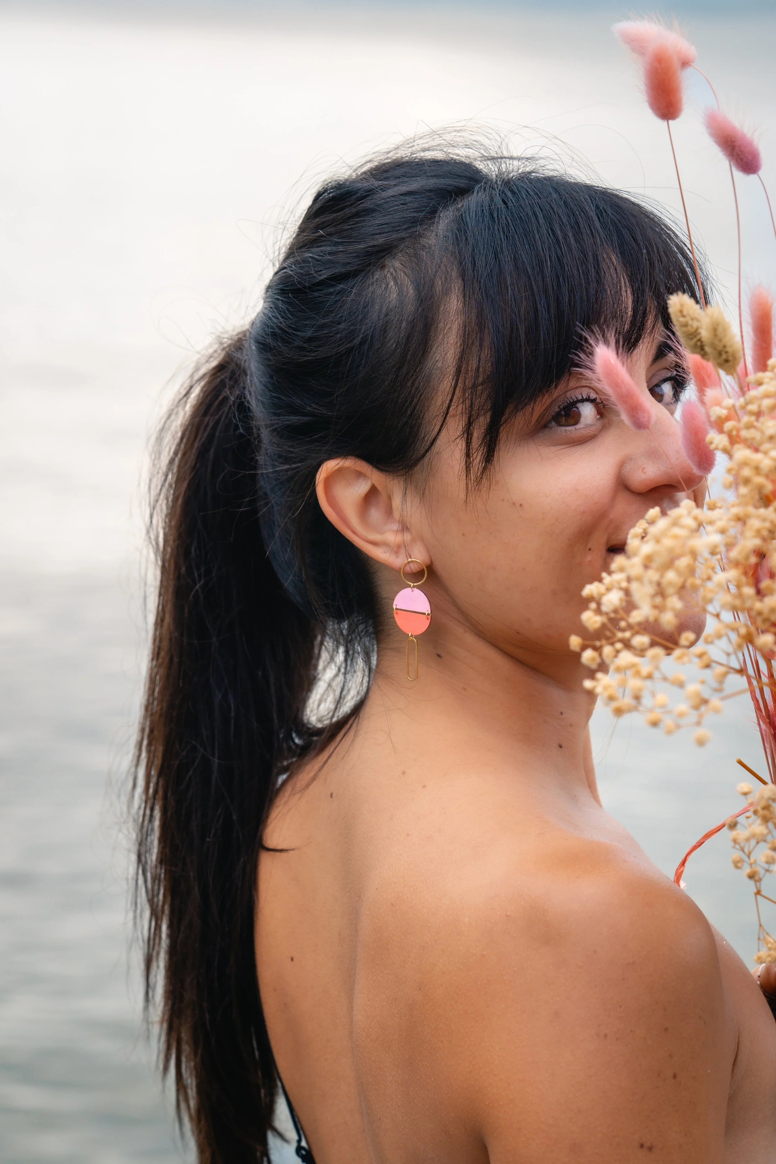 Femme aux cheveux noirs, portant une boucles d'oreilles colorées, se tenant près d'un lac ou d'une mer, tenant un bouquet de fleurs pastel.