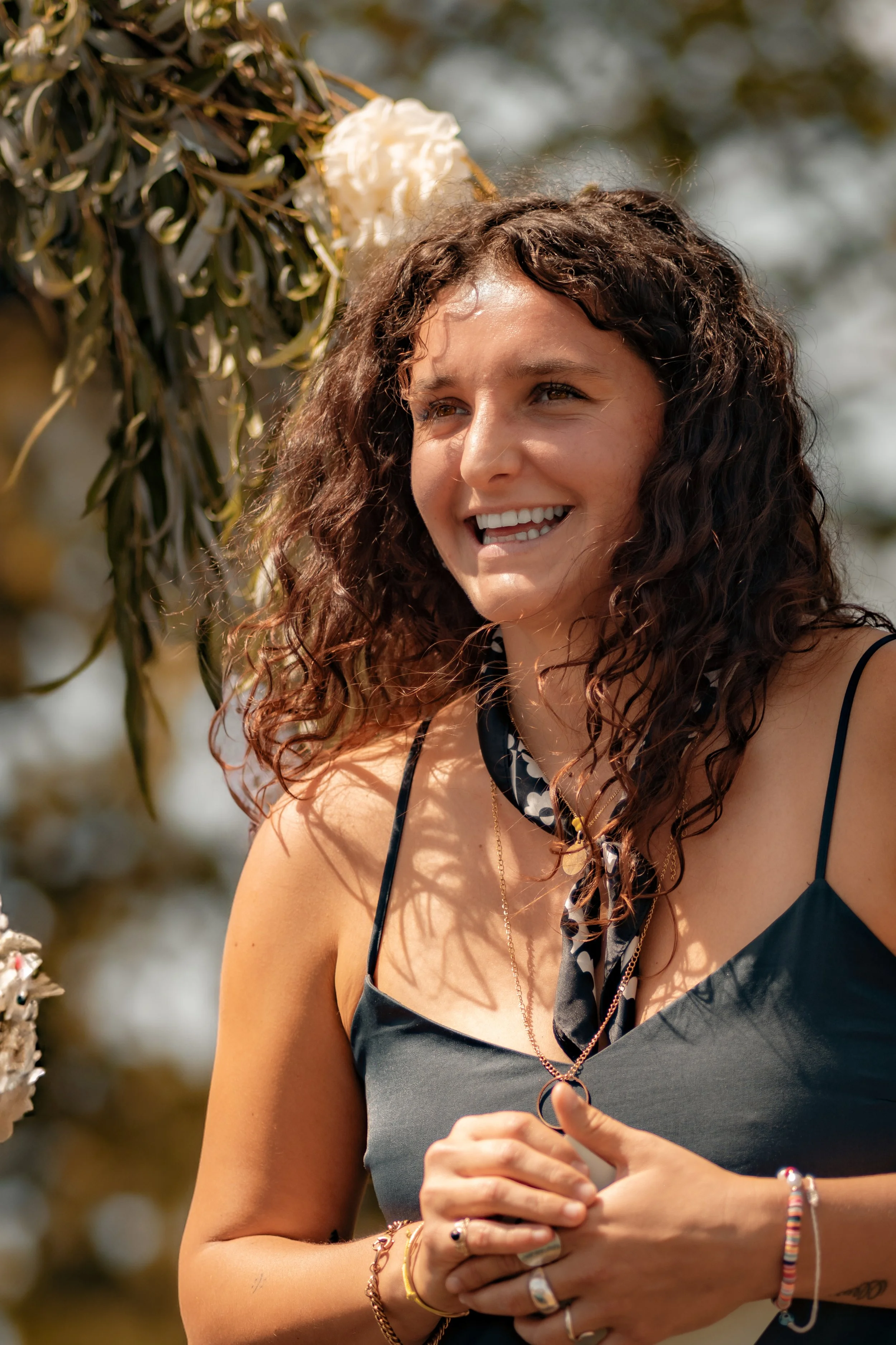 Une femme souriante avec des cheveux bouclés foncés portant une robe noire et un foulard à pois, avec des bijoux, lors d'une cérémonie en extérieur.