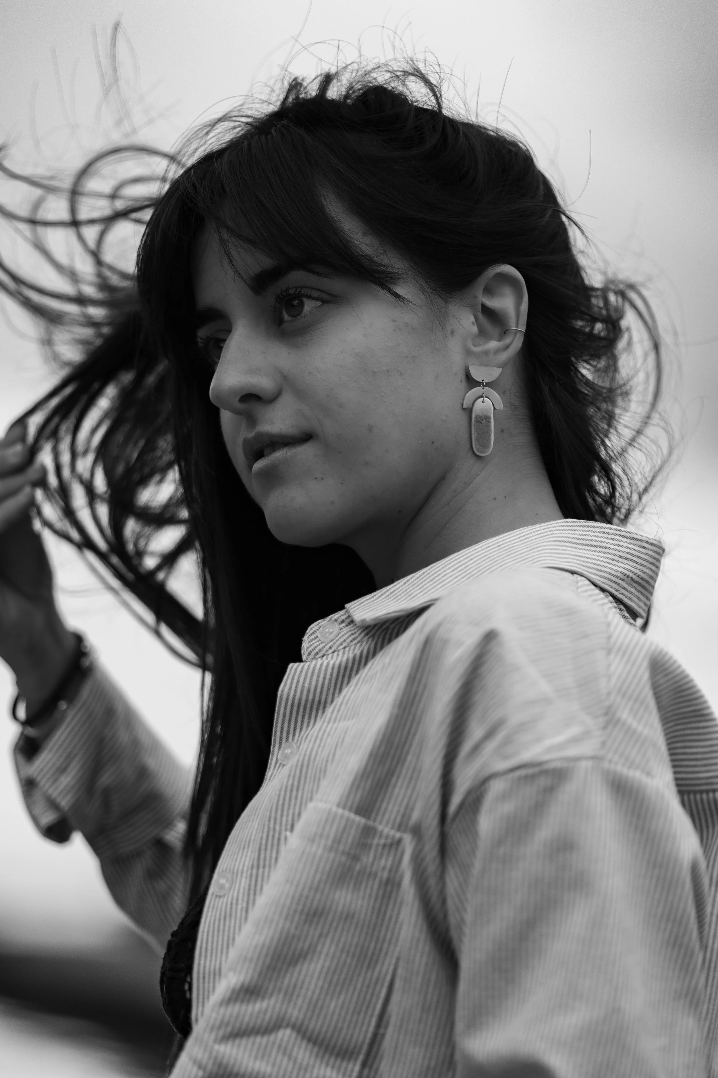 Portrait en noir et blanc d'une femme aux cheveux longs qui bougent, portant des boucles d'oreilles modernes et une chemise à rayures, regardant vers la gauche.