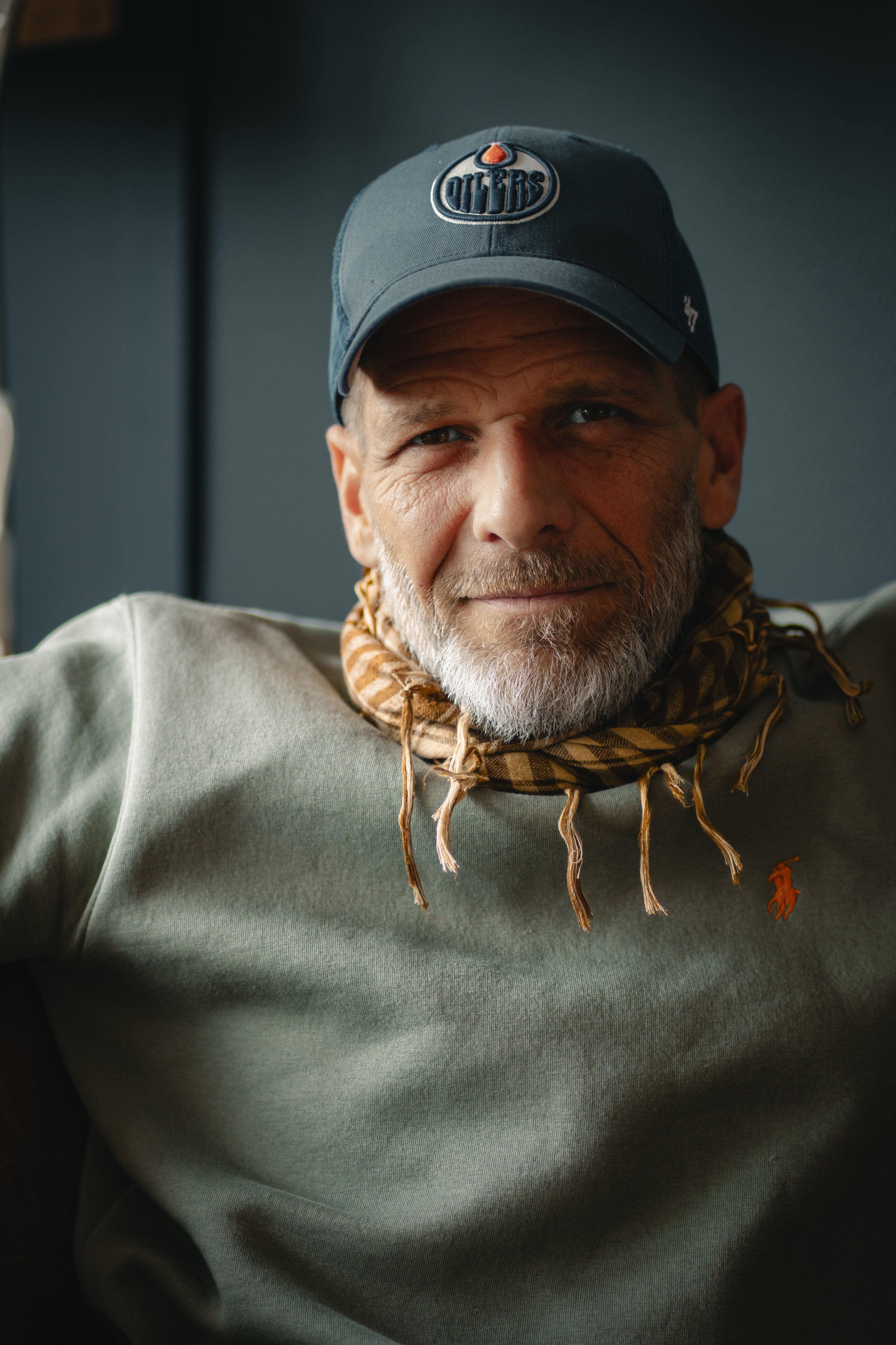 Un homme avec une barbe grise portant une casquette bleue, un pull gris et une écharpe à carreaux, assis dans un endroit sombre.
