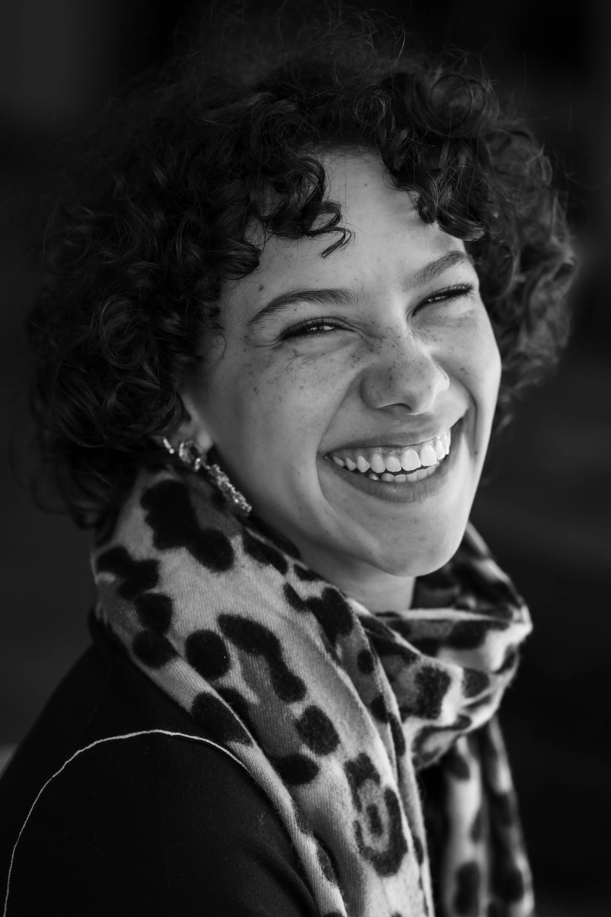 Une femme souriante avec des cheveux bouclés courts, portant un foulard à motifs léopard, des boucles d'oreilles et un sourire éclatant.