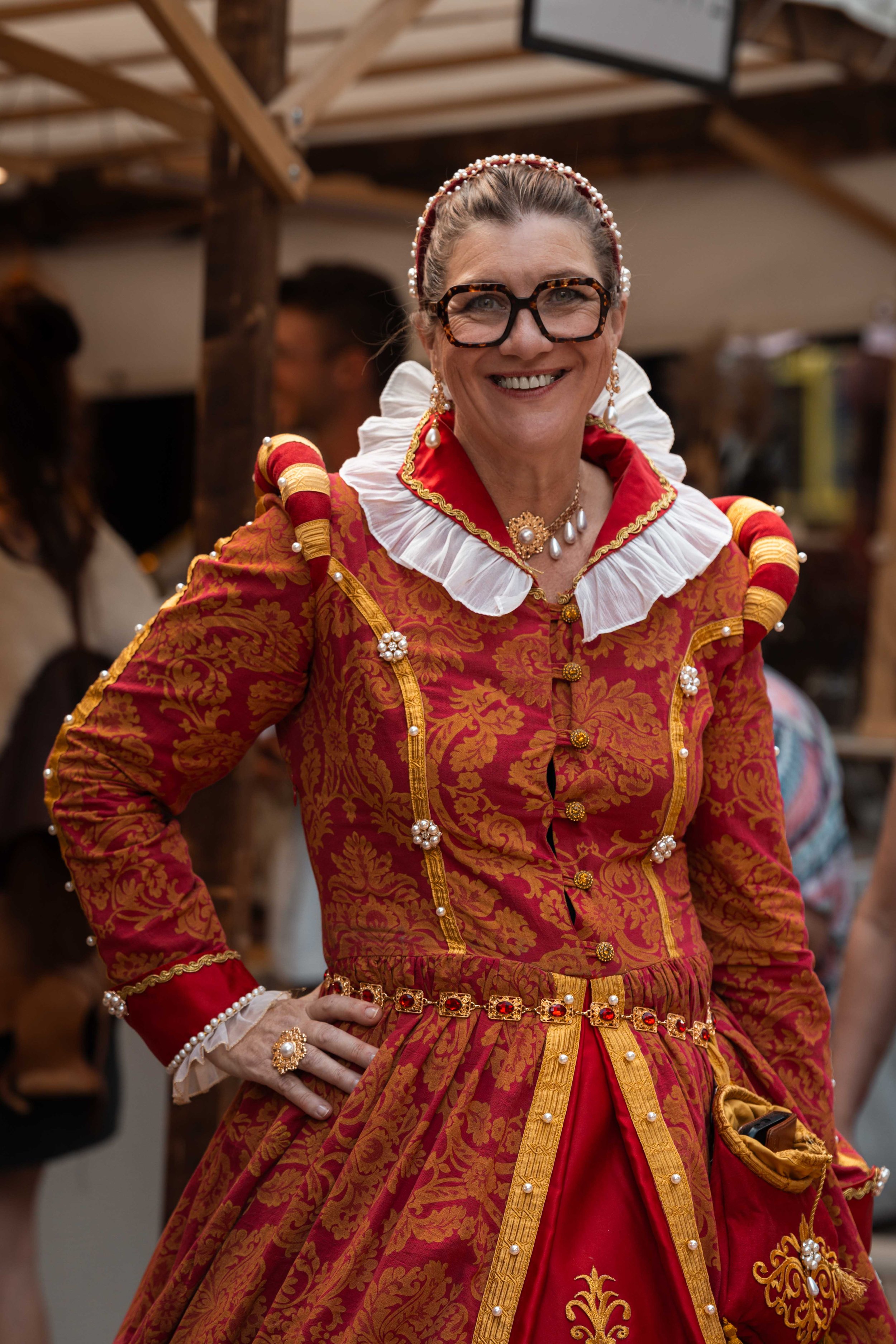 Femme souriante portant un costume traditionnel richement décoré en rouge et or avec des accessoires de style ancien, portant des lunettes.
