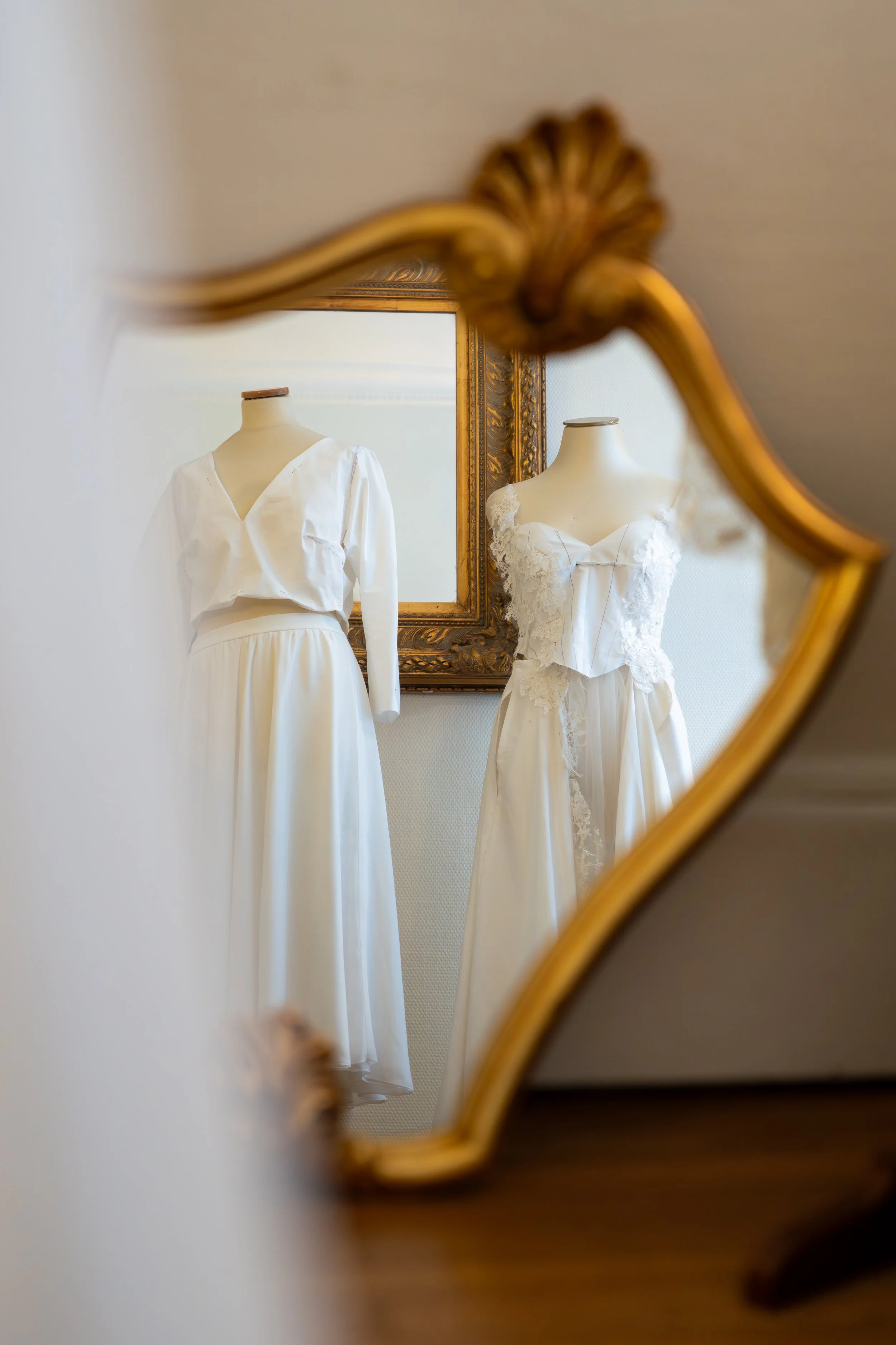 Deux robes de mariée blanches affichées devant un miroir orné d'un cadre doré, vue à travers un miroir antique avec un cadre en bois décoratif.