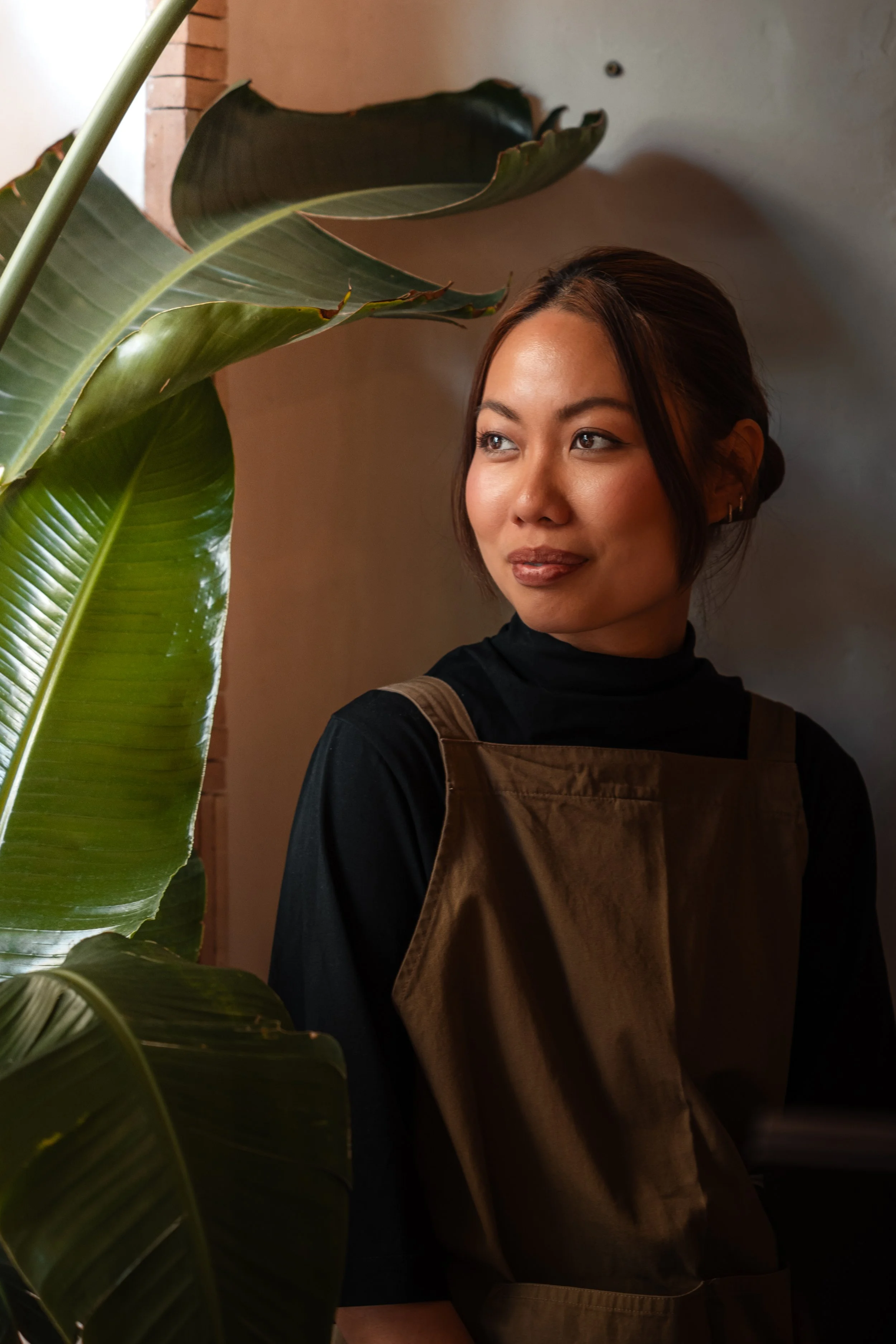 Une femme souriante portant un tablier marron, debout près d'une grande plante verte, dans un intérieur avec mur gris et décor en briques orange.