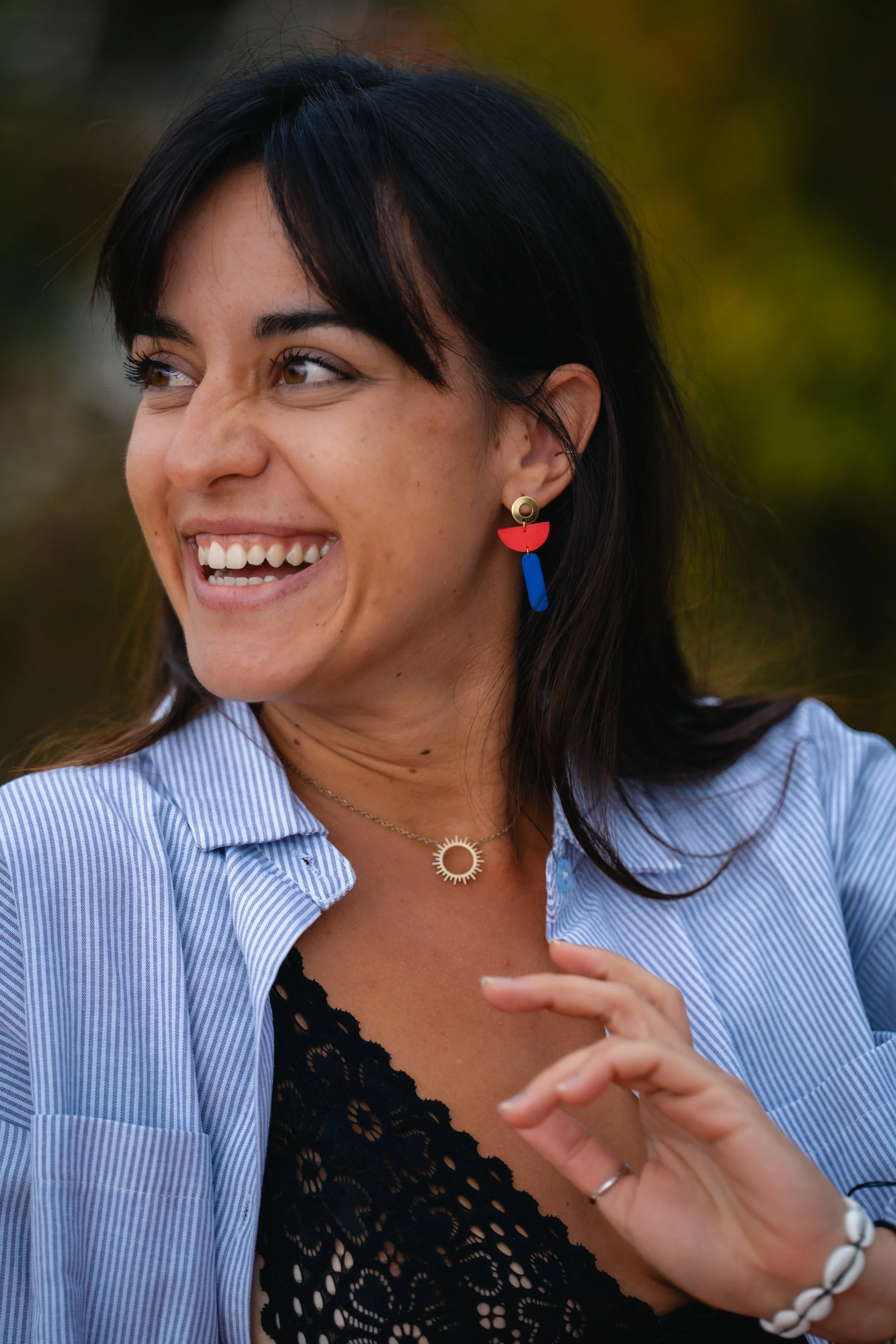 Jeune femme souriante avec boucle d'oreille colorée, portant un collier en forme de soleil, vêtue d'une chemise à rayures bleues et d'une robe en dentelle noire, avec un fond flou de nature.