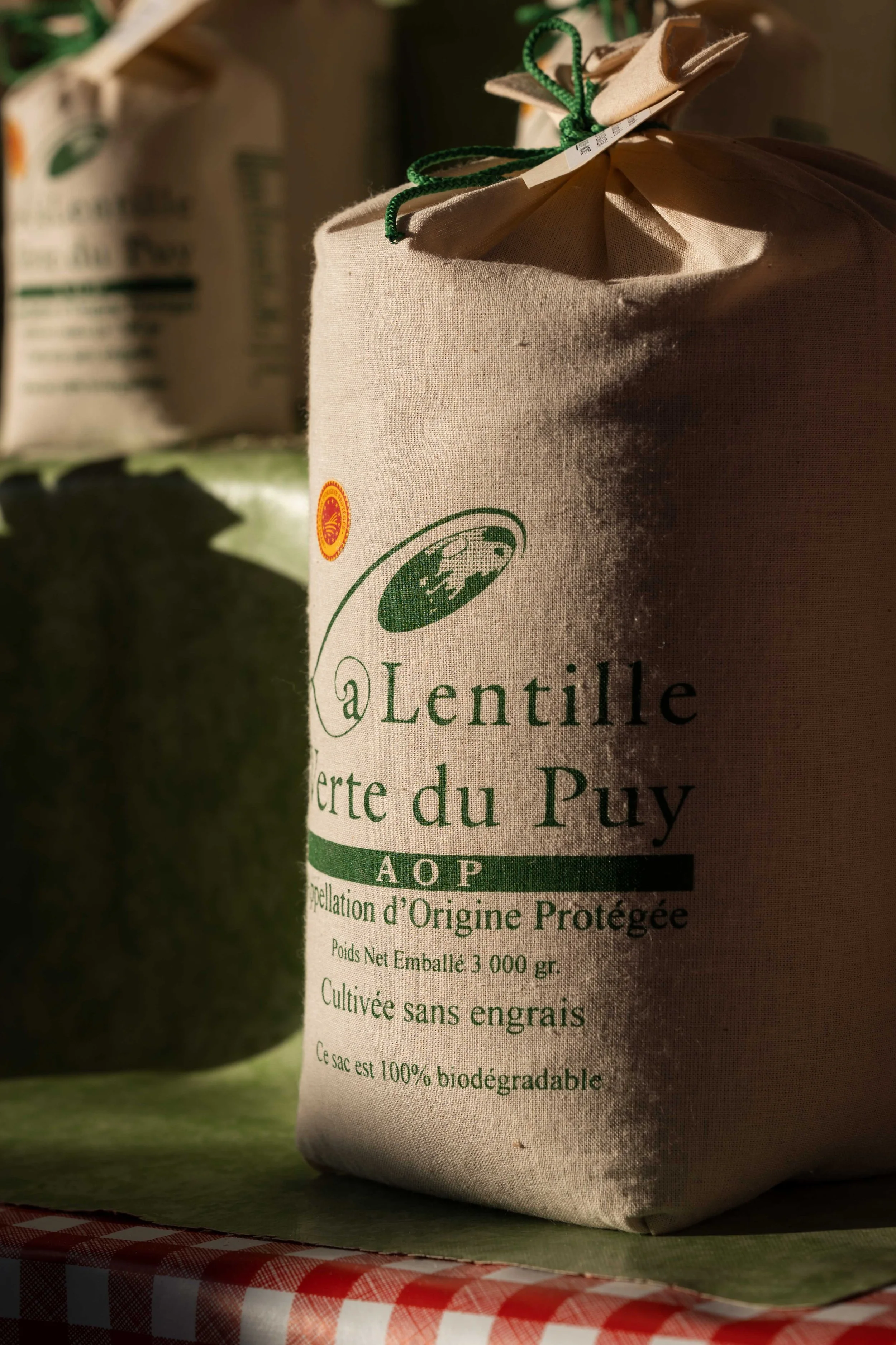 Sac en toile beige avec inscription en français, mentionnant l'origine protégée, cultivé sans engrais, biodégradable, contenant 3000 grammes de lentilles vertes du Puy, posé sur une nappe à carreaux rouges et blancs, avec d'autres sacs de lentilles e