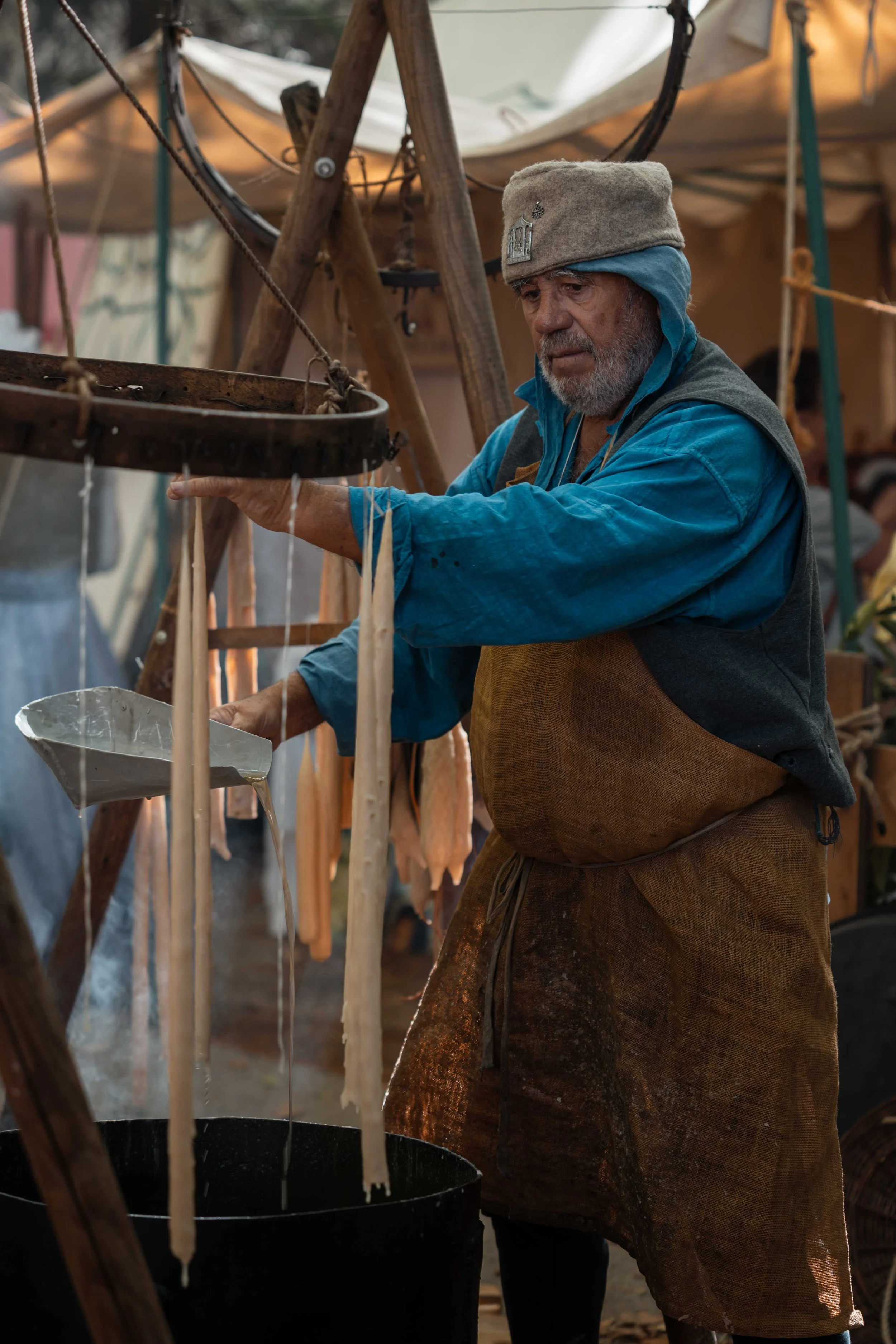 Un homme âgé, portant un chapeau en laine et des vêtements amples, en train de préparer du fromage dans un marché ou une ferme. Il verse du liquide blanc d'une bassine dans un récipient, avec un décor en bois et des tents en toile en arrière-plan.