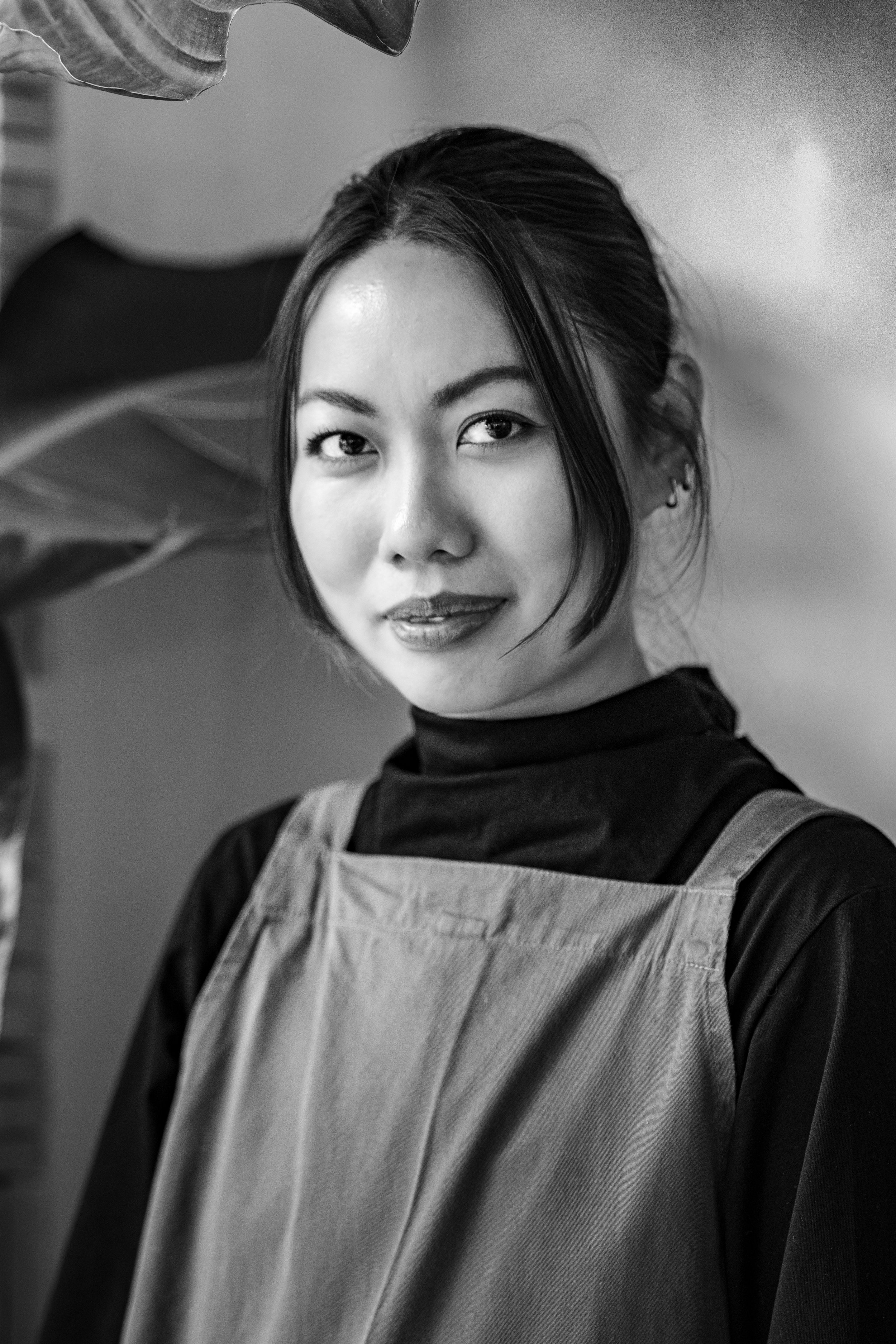 Portrait d'une femme asiatique portant un tablier sur un haut noir, souriante légèrement, avec un espace flou en arrière-plan.
