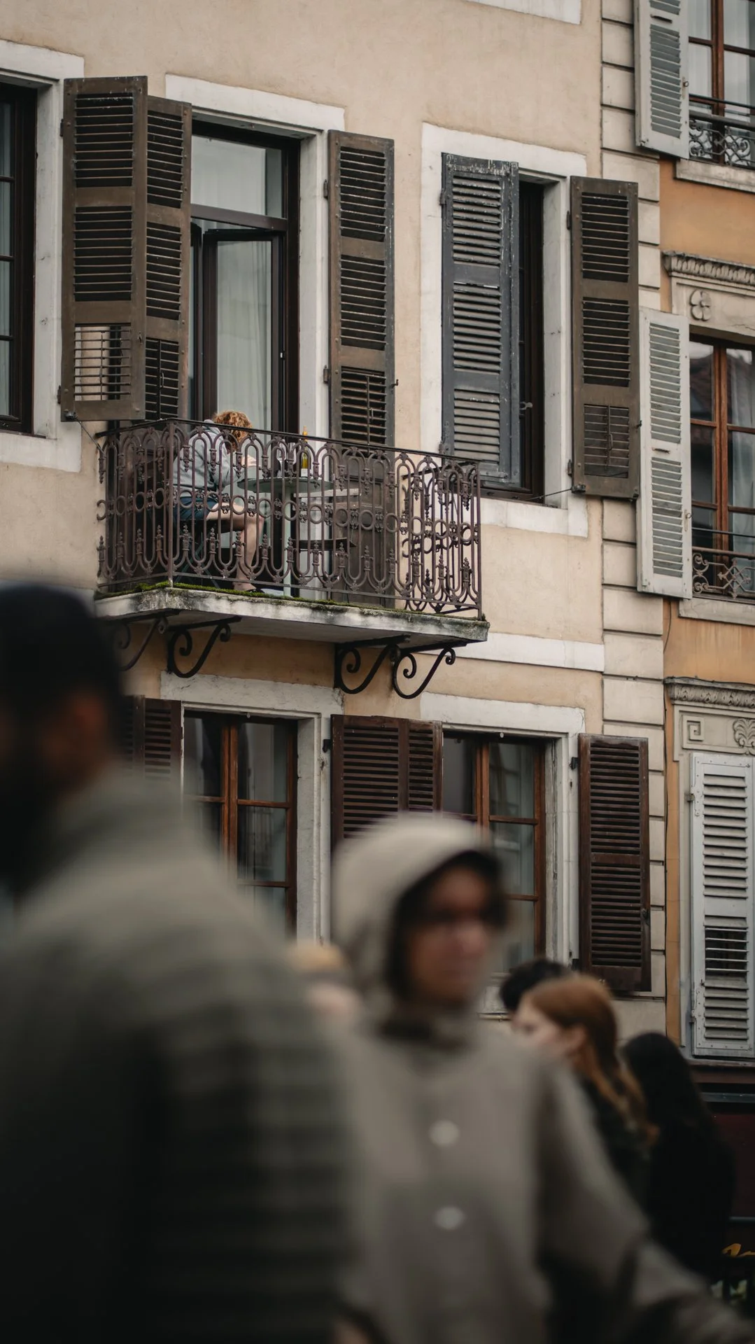 Une femme assise sur un balcon en face d'un bâtiment avec des volets en bois. Plusieurs personnes marchent au premier plan, floues, dans une rue urbaine.