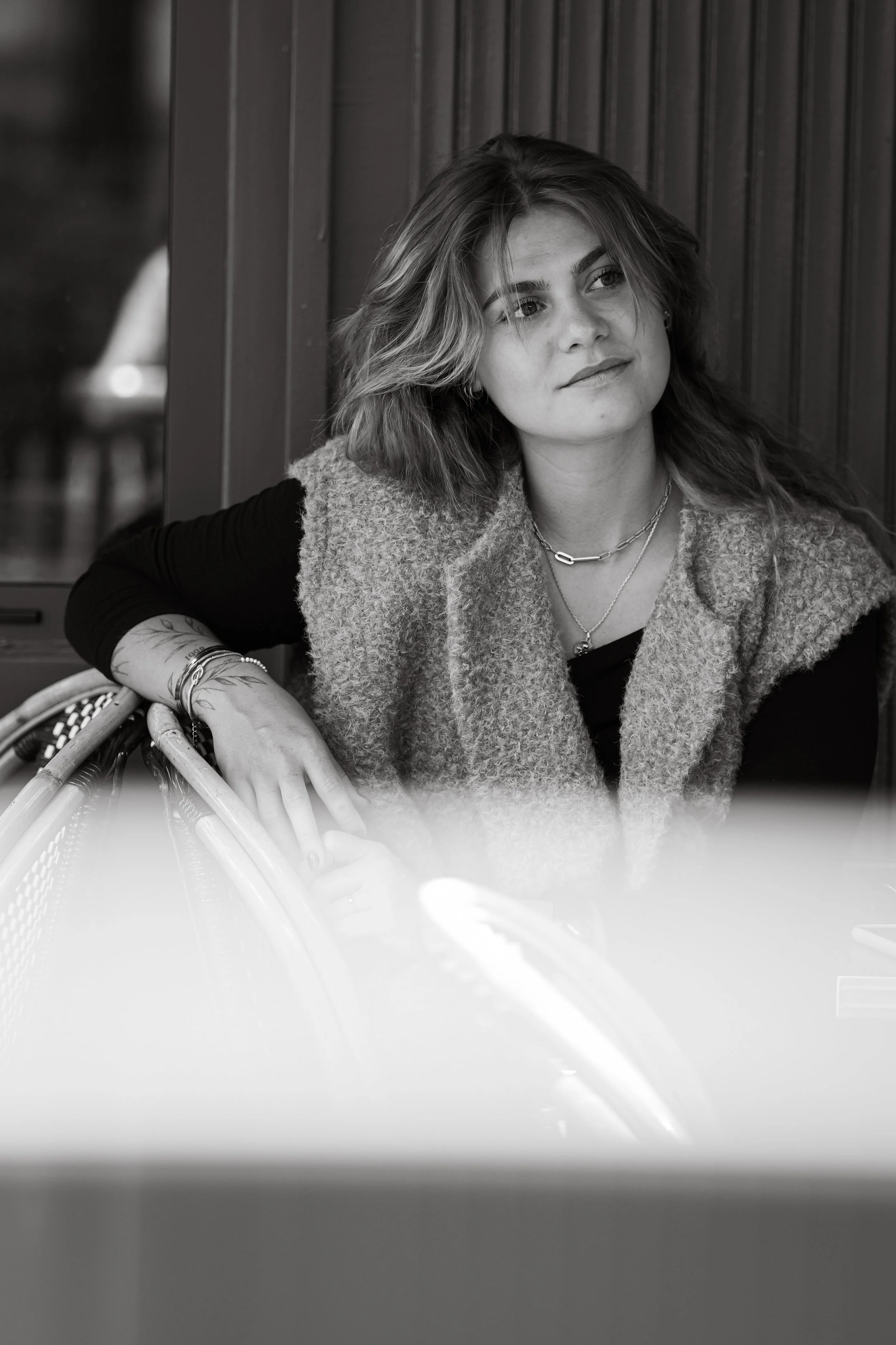 Jeune femme assise dans un café, regardant pensivement vers la droite, avec des cheveux ondulés, portant des bijoux et une veste en laine over size, en noir et blanc.