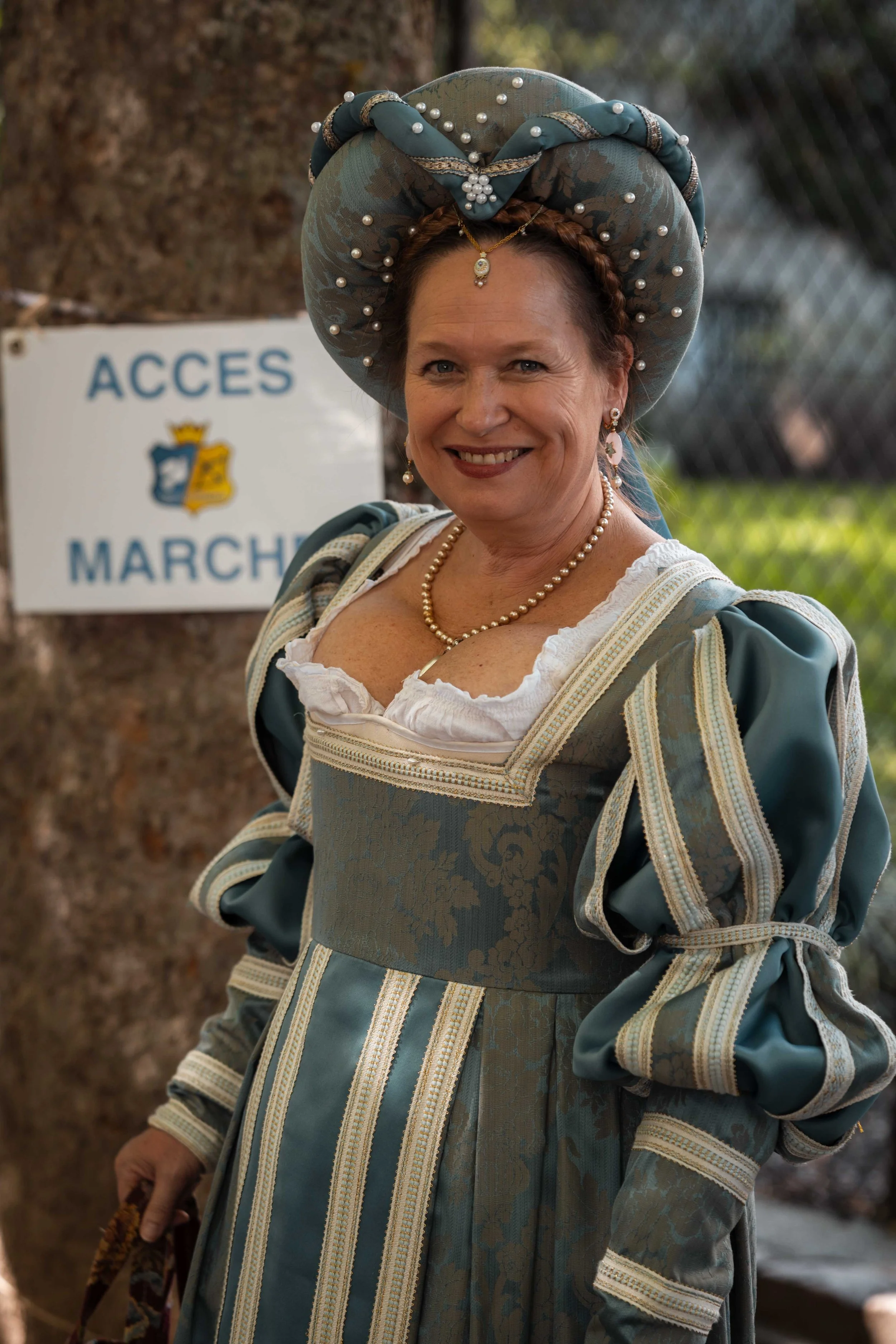 Femme en costume d'époque avec un grand chapeau décoré de perles, souriante, portant un collier de perles et une robe ornée de motifs, en extérieur près d'un panneau 'Accès Marché'.