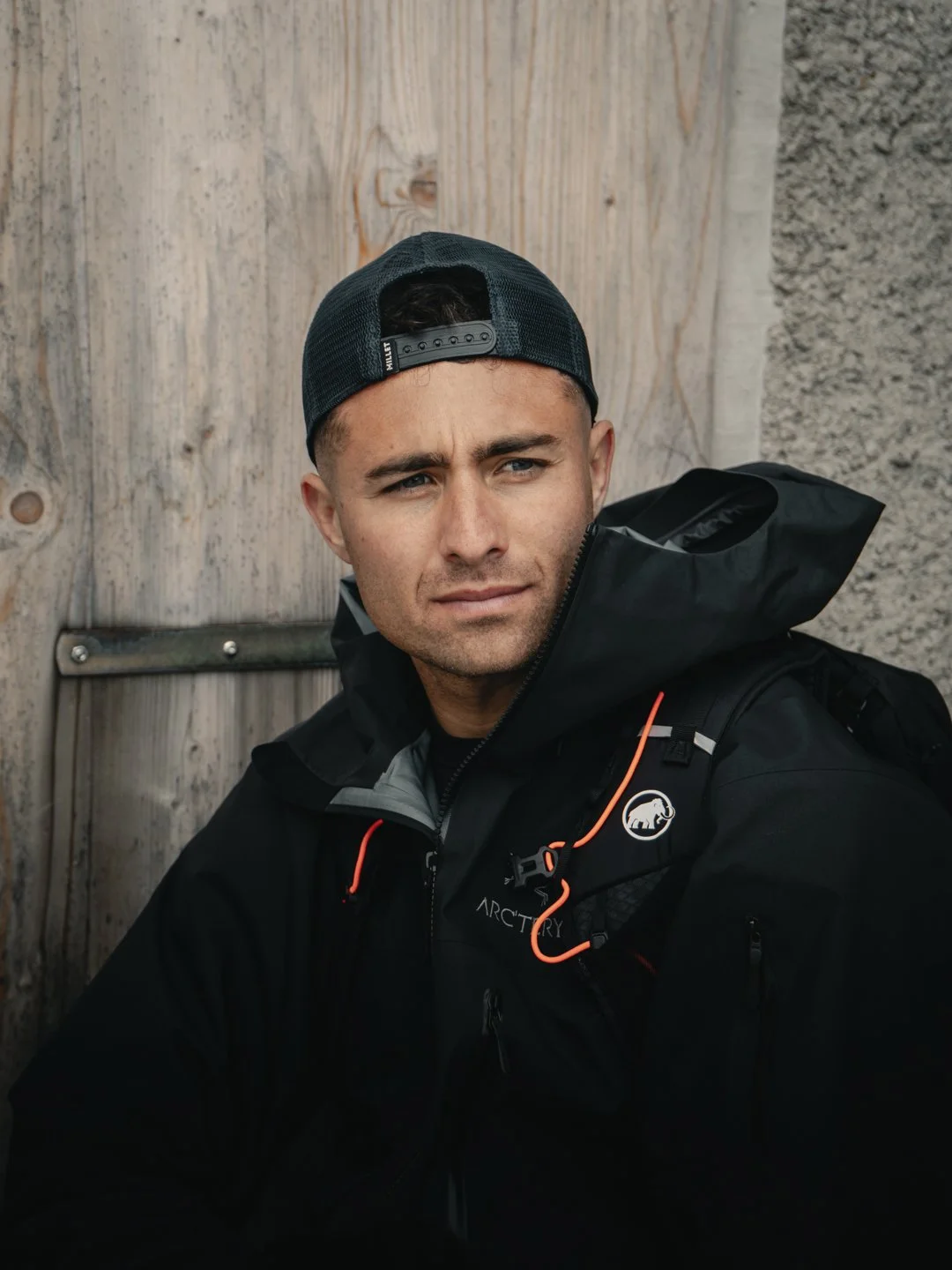 Un homme portant une casquette noire et une veste noire avec un logo Arcteryx, assis contre un mur en bois et en pierre.