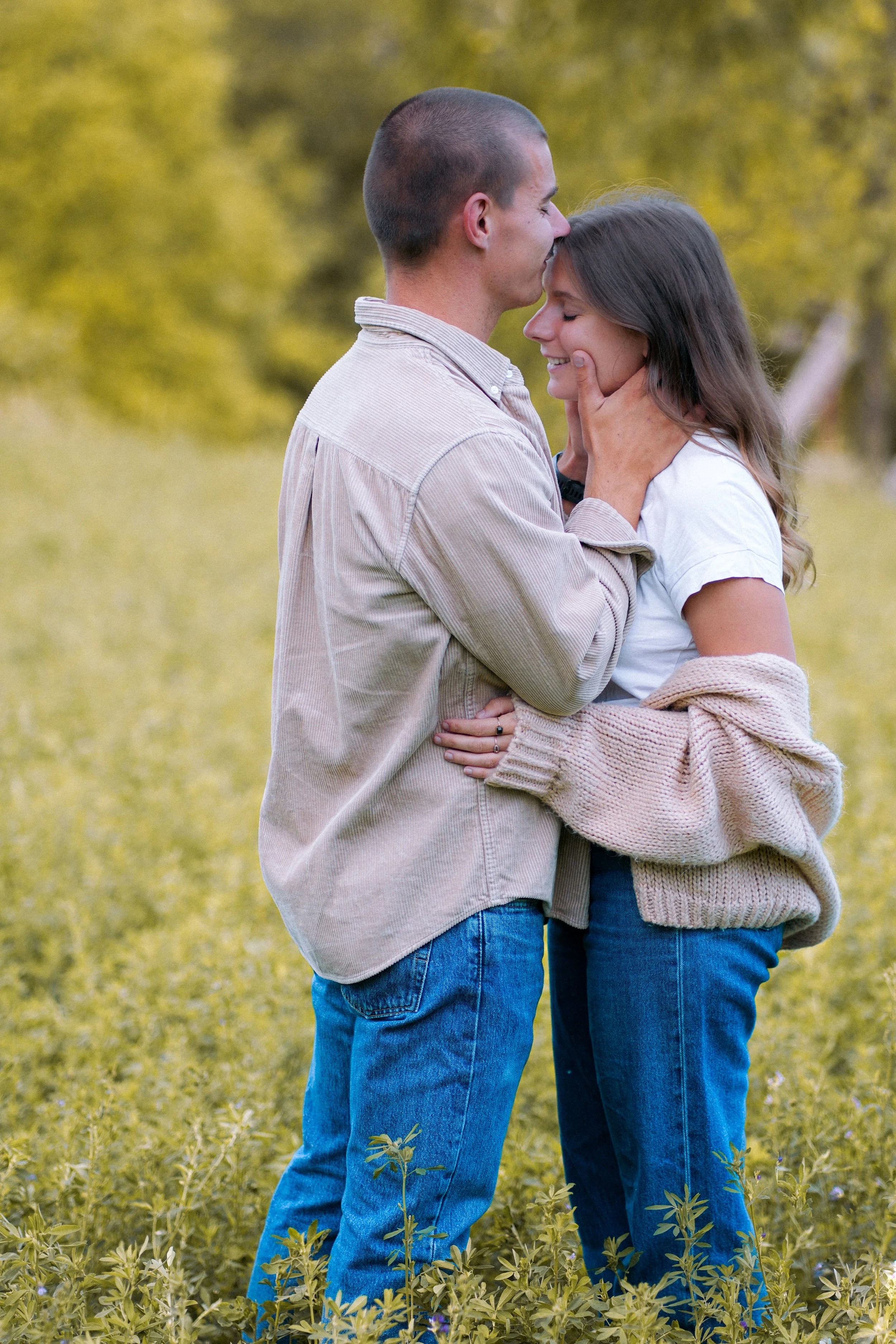 Un couple enlacé dans un champ, échangeant un baiser, avec arrière-plan de feuillage automnal.