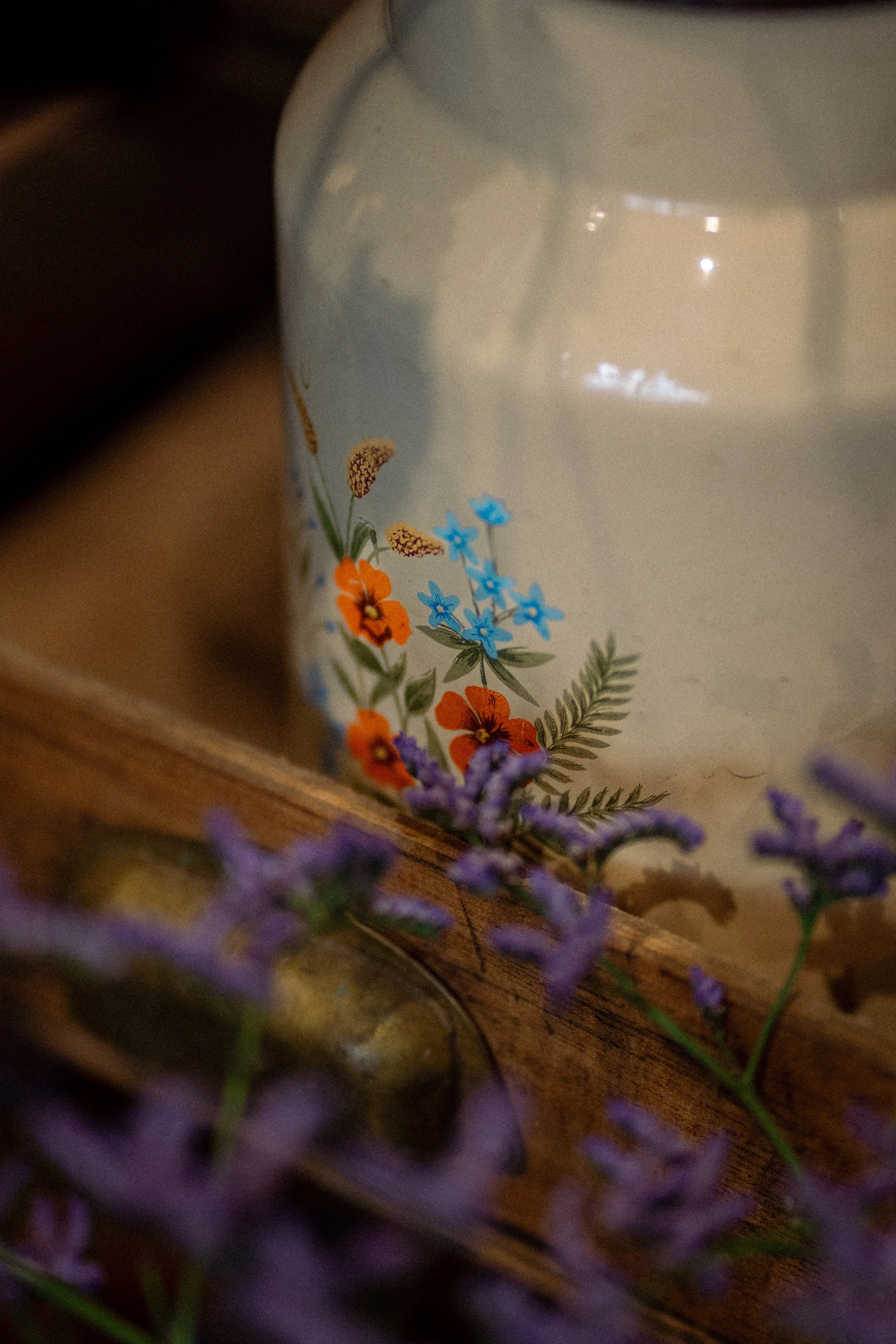 Un pot en céramique blanc avec un motif floral coloré, placé sur une surface en bois, entouré de fleurs de lavande violettes.